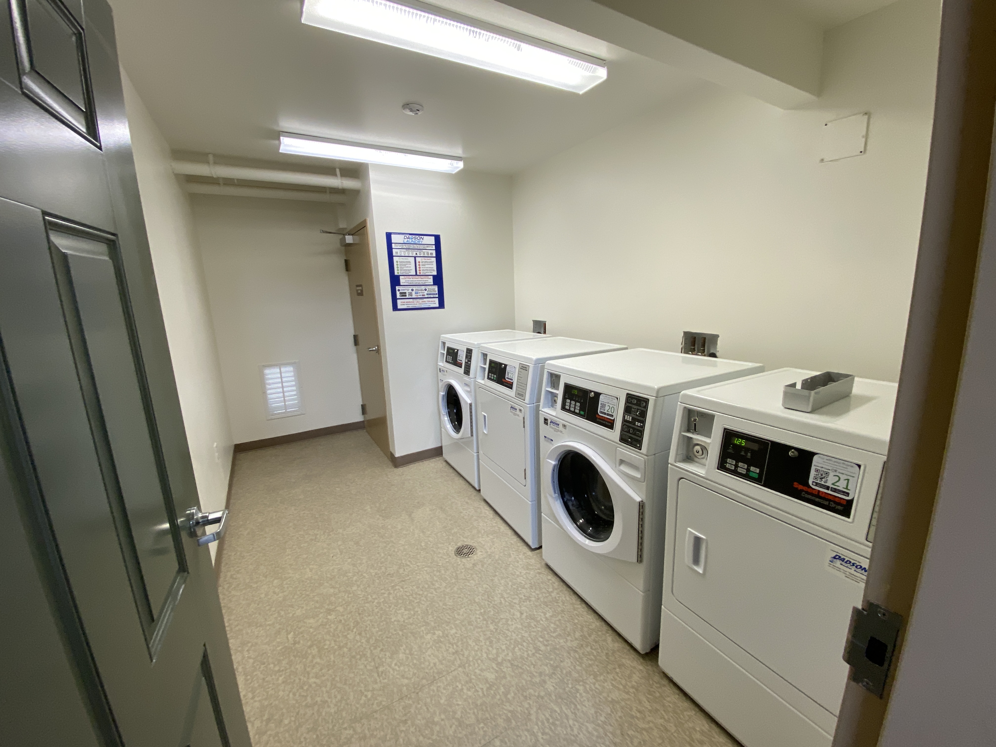 Laundry room Machines line the wall. Blue poster on the far wall. Next to a brown door.