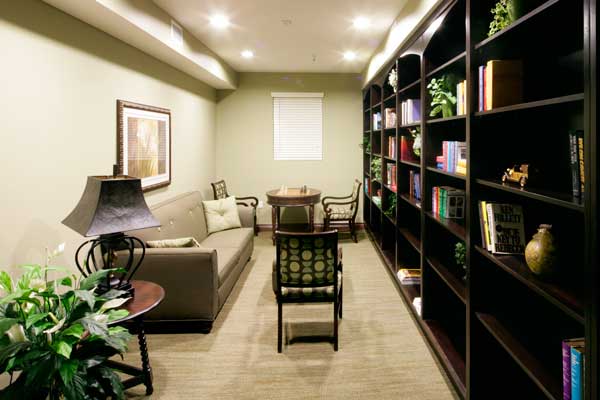 Interior image of Andalucia Senior Apartmens library. A dark brown book case along one entire wall, with round chess table, chairs, couch and side table with a lamp and plant on it.