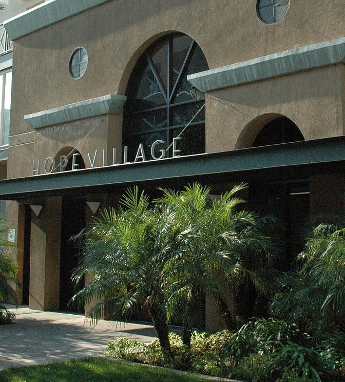 Front angle view of a light brown building. There is a grass area, trees and plants on the side leading to the entrance. Above the entrance the header reads, "Hope Village".