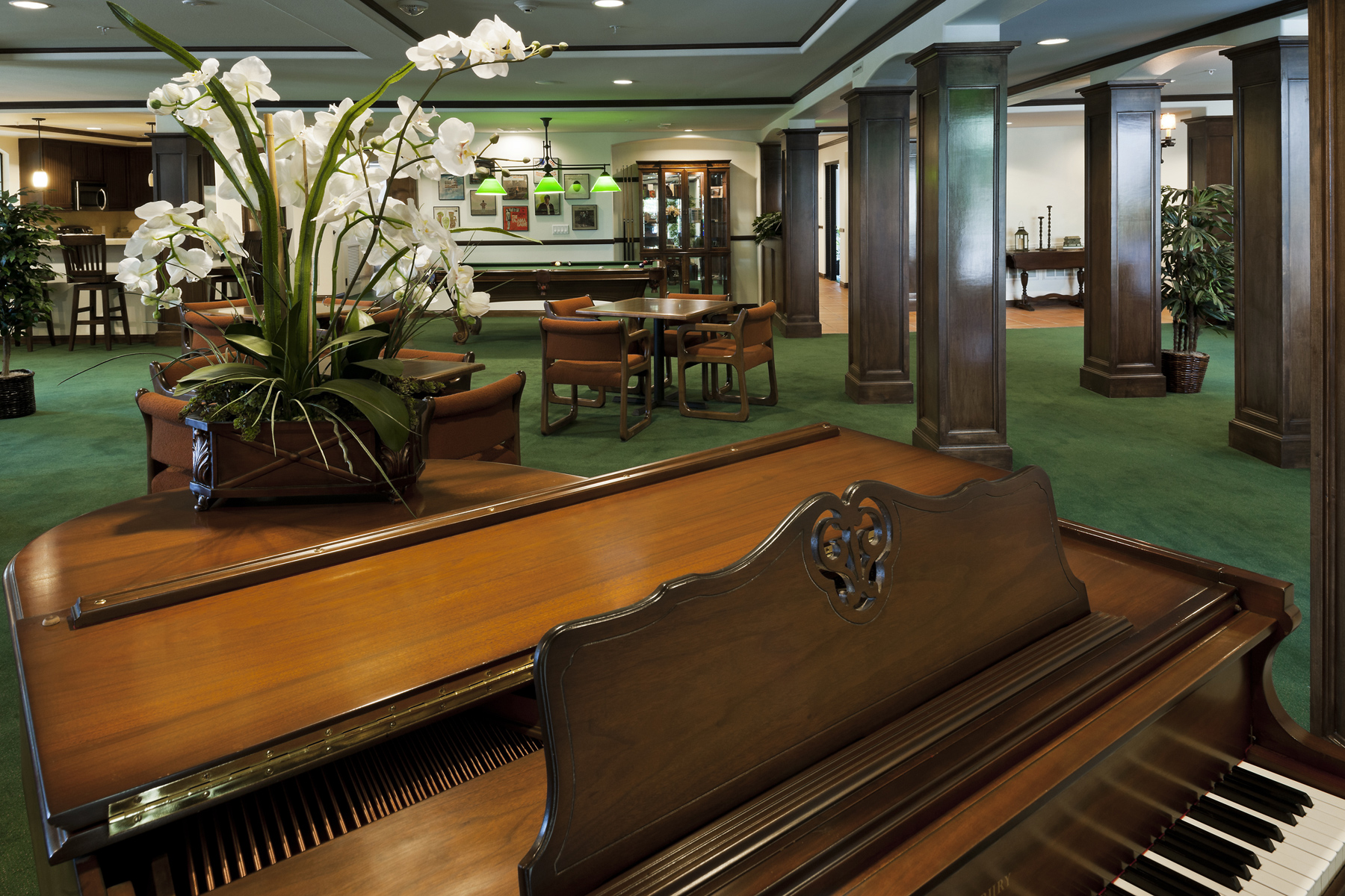 Canby woods housing community room. room wood with green carpet, several brown wood seating and tables, grand piano, plants throughout room, painting on wall. Large pillars and recessed lighting