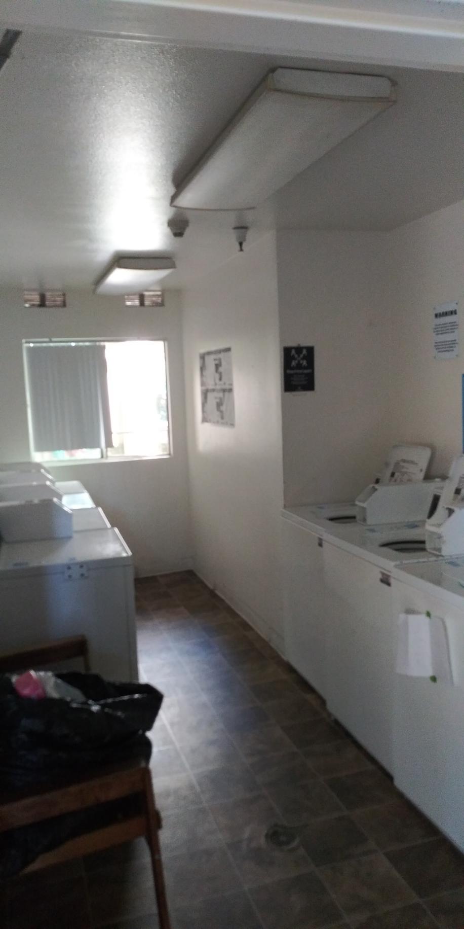 Laundry room with six machines acroos from each other. There is a window with blinds, and the floor is tiled.