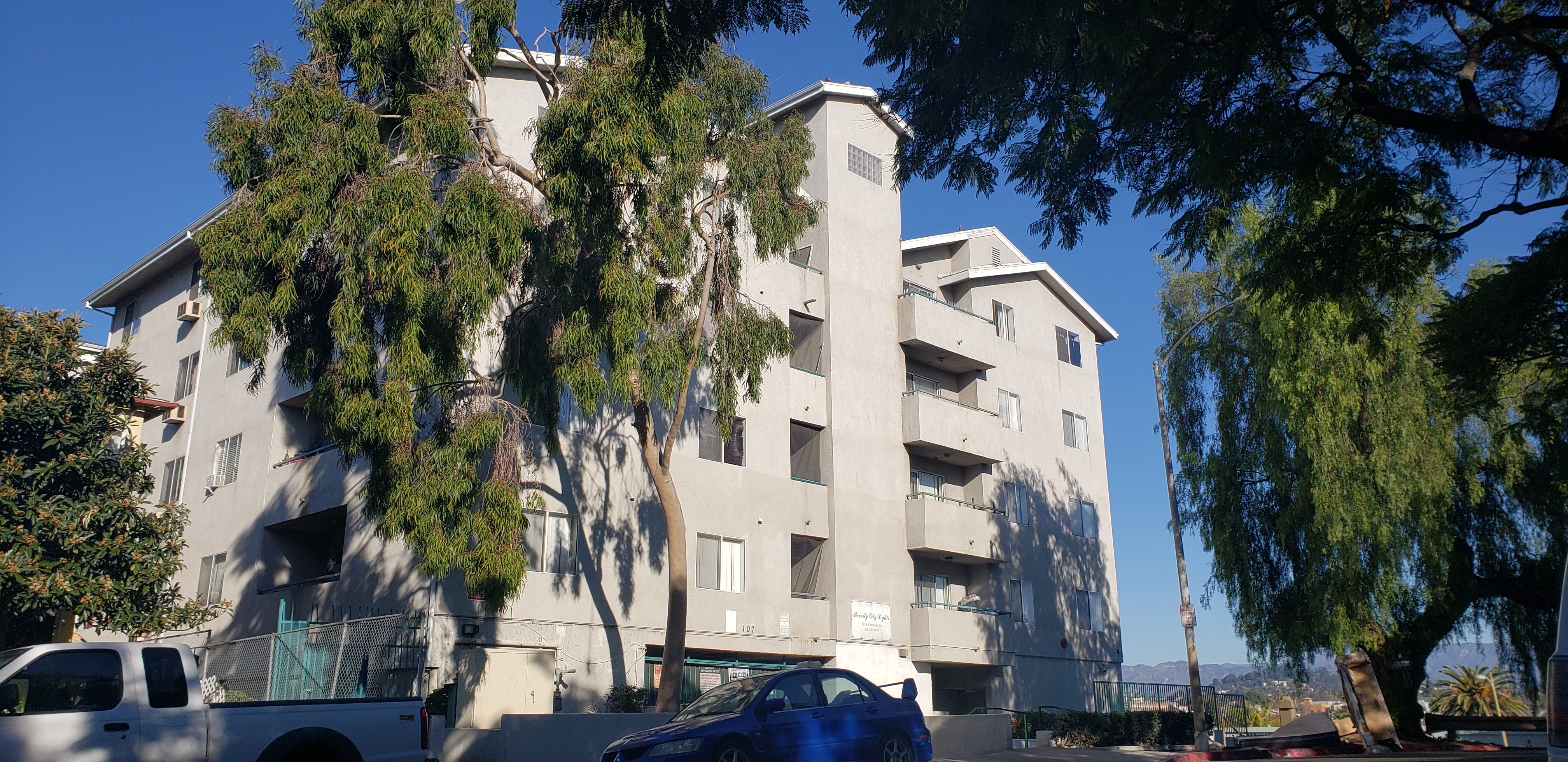 Front view of Beverly city lights. Multi-level light grey building. Units with balcony access. Parking garage gate located at bottom of building. Building entrance has stairs leading down. Street and sidewalks lined with trees and bushes. Cars parked on s