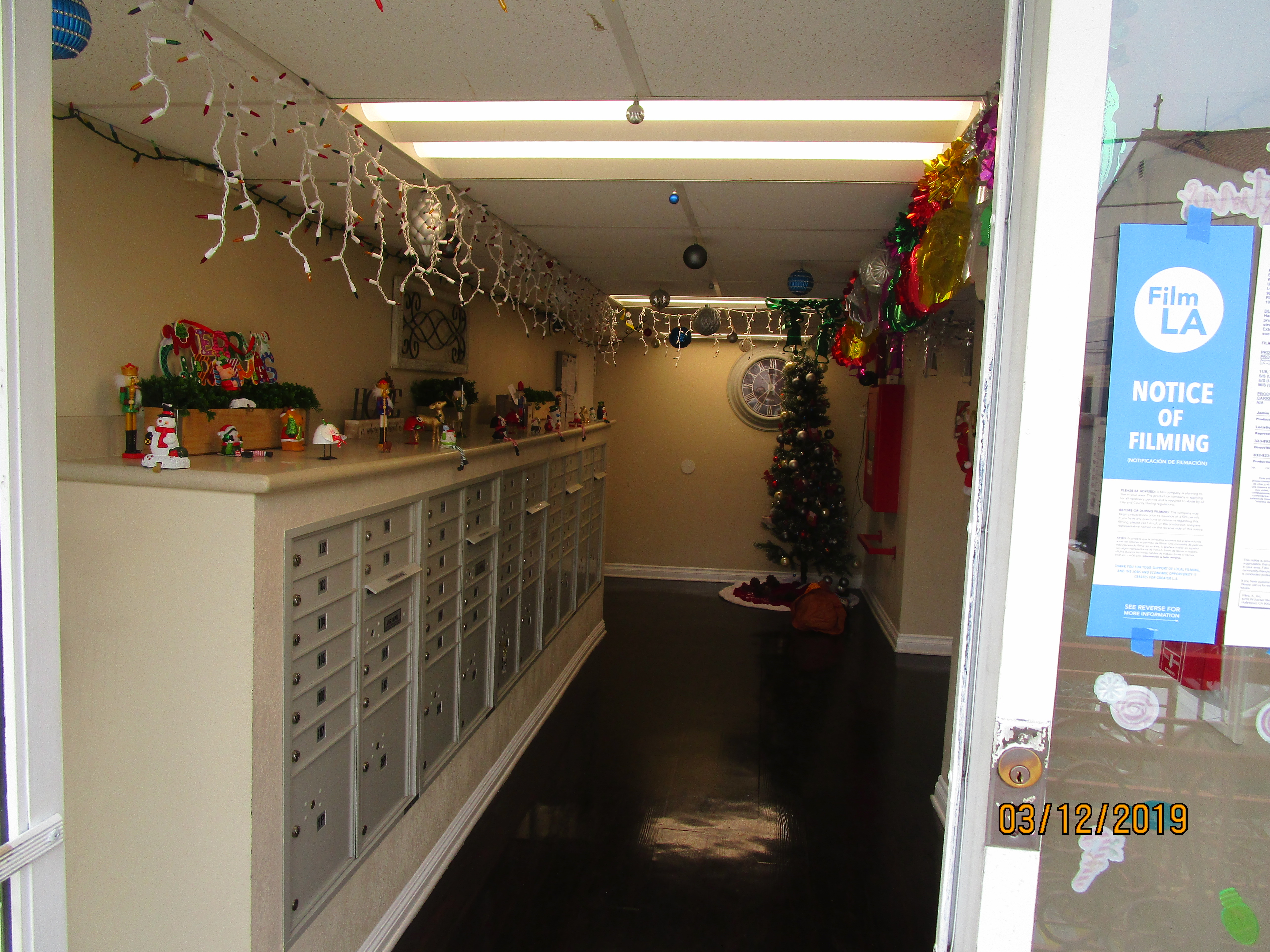 View of a mail room, lights and Christmas ornaments hanging from the office-like ceiling, ornaments on top of the mailboxes shelve, A christmas tree on the right side corner at the end of the room, a big decorative clock next to the tree, a notice of film