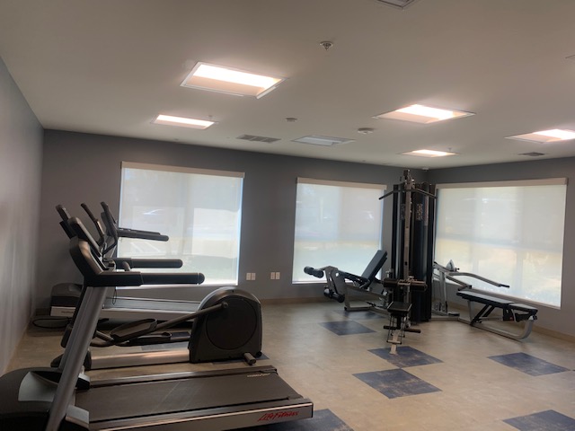 View of a small gym, workout equipment, three windows with white shades, linoleum floors.