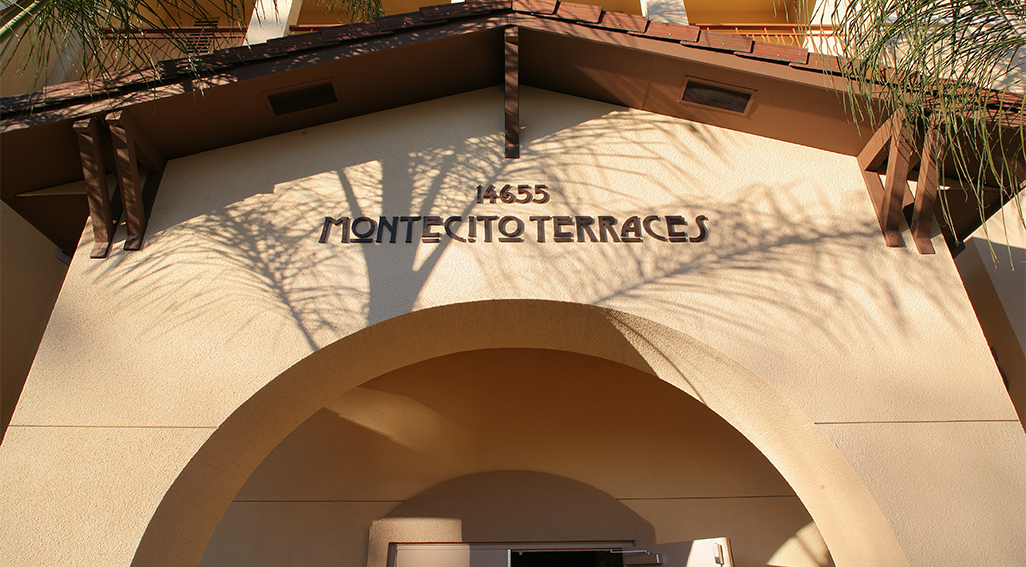 View of the overhead of a peach-colored entrance. It reads "Montecito Terraces" in Art Nouveau letters and has a hut shaped top, with two king palms just inside the frame.