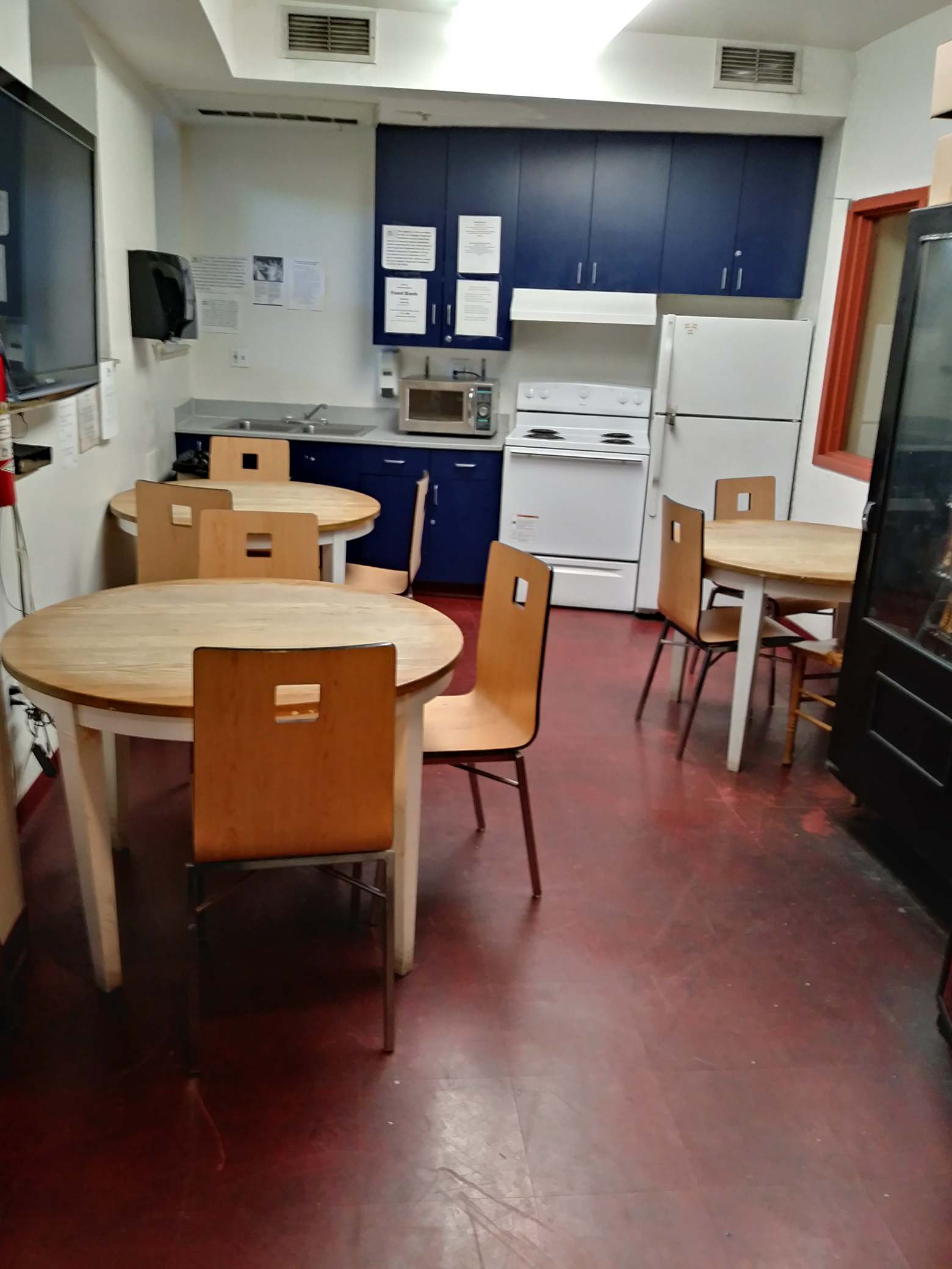 Photo of Community Kitchen Area showing three round tables with chairs, kitchen cabinets, refrigerator, stove and oven, microwave and flat screen TV attached to the wall.