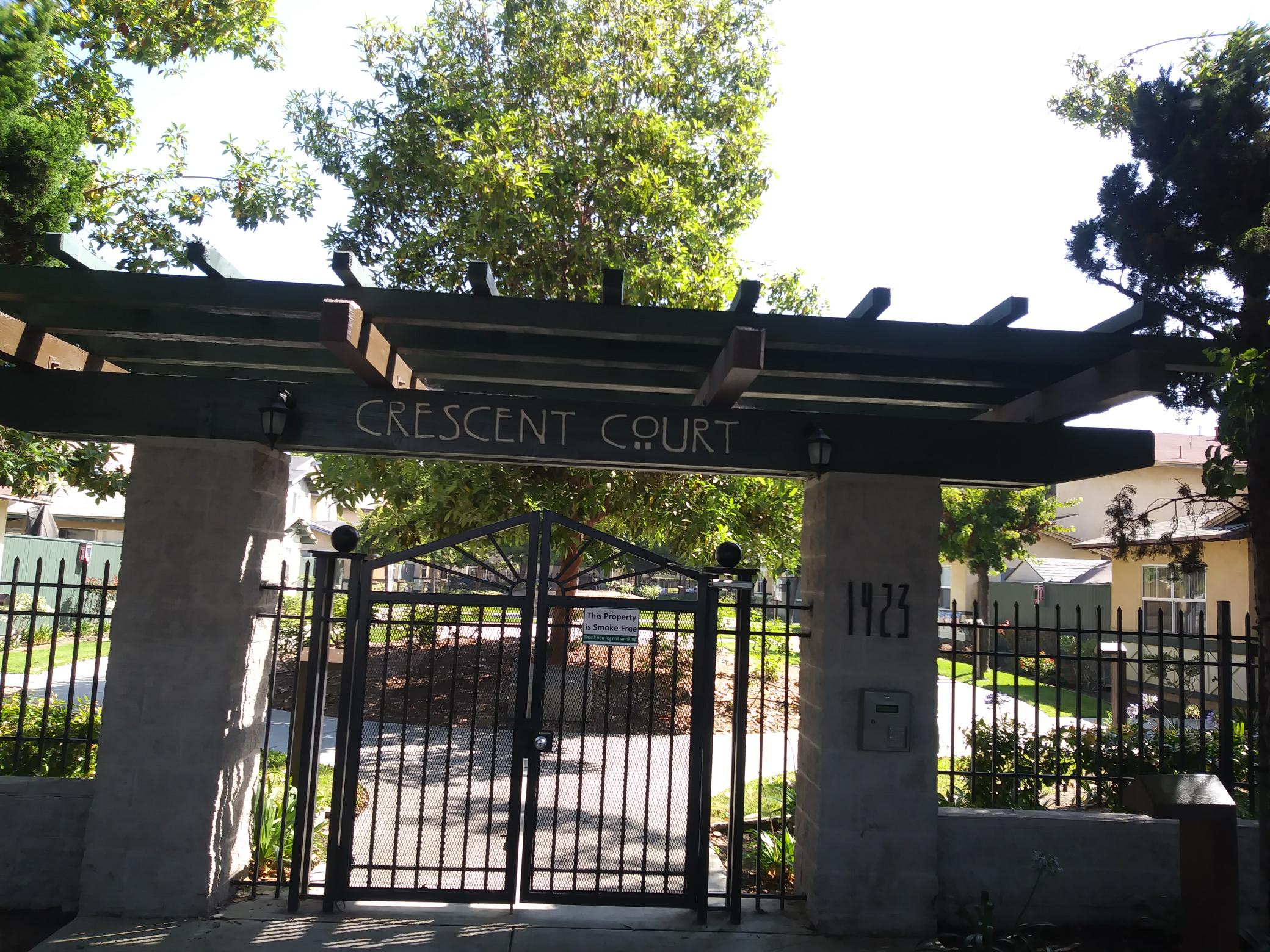 Front view of Crescent Court Apartments. Large gate leading into courtyard. Large tree in the center and on either side of it there is side walk access. Along walkway is grass and flower beds.
