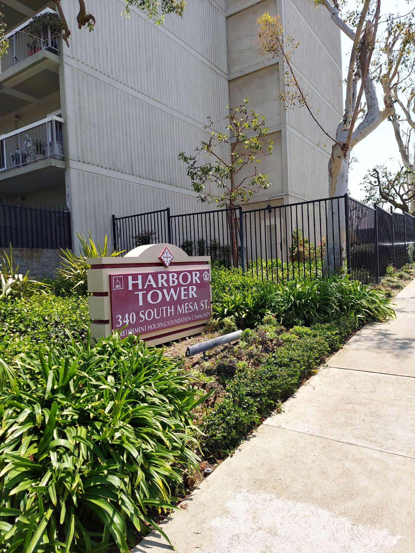 Plant area in front of a building. There is a sign in the middle that reads "Harbor Tower" and a fence in the back that separates this section with another plant section.