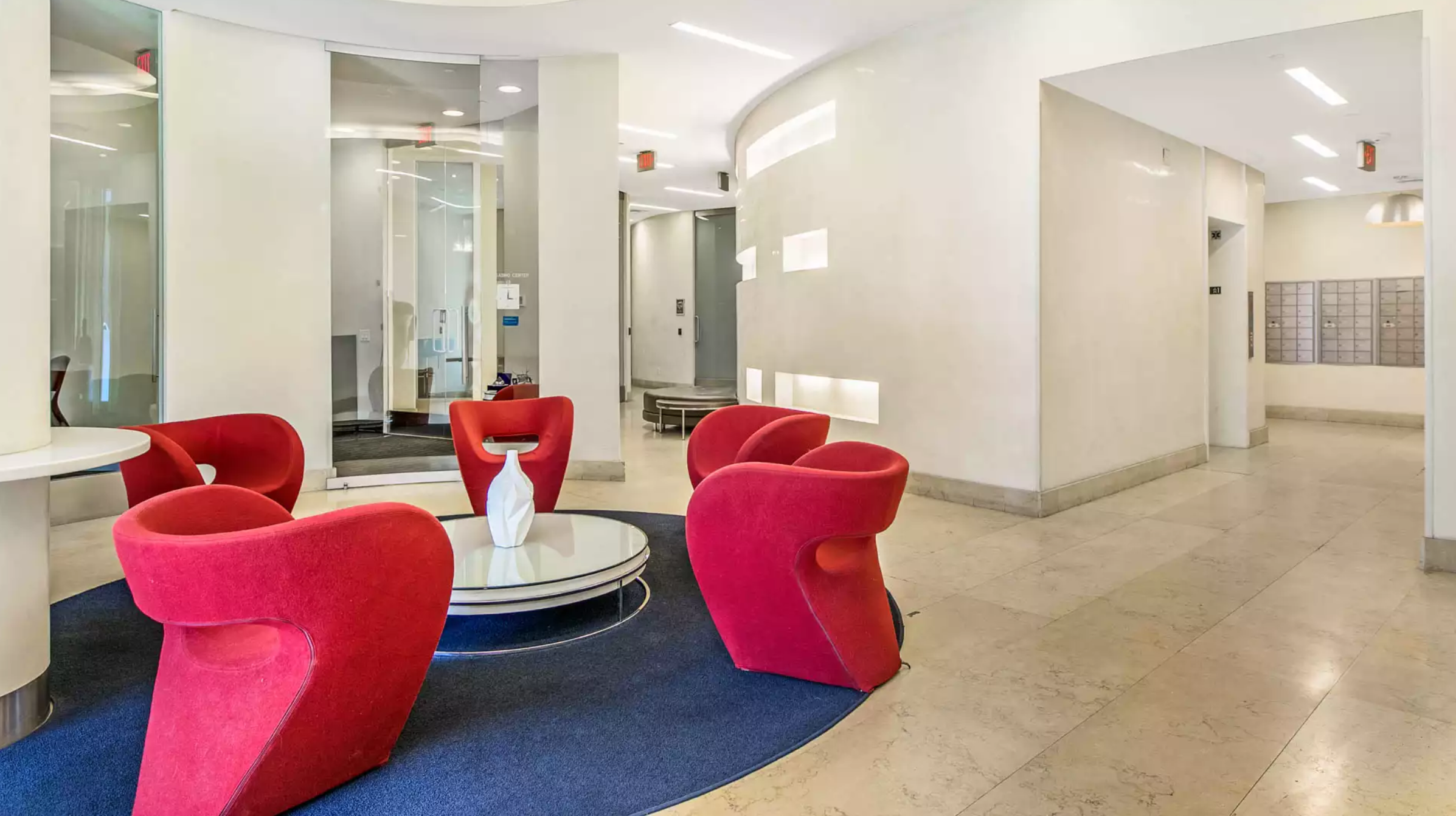 Inside view of a lobby area. There are modern sofa chairs that surround a small round table. To the right side there is an elevator entrance and a mailbox area. To the left is a curbed hallway and tall windows from another room. Soace is well litted all a