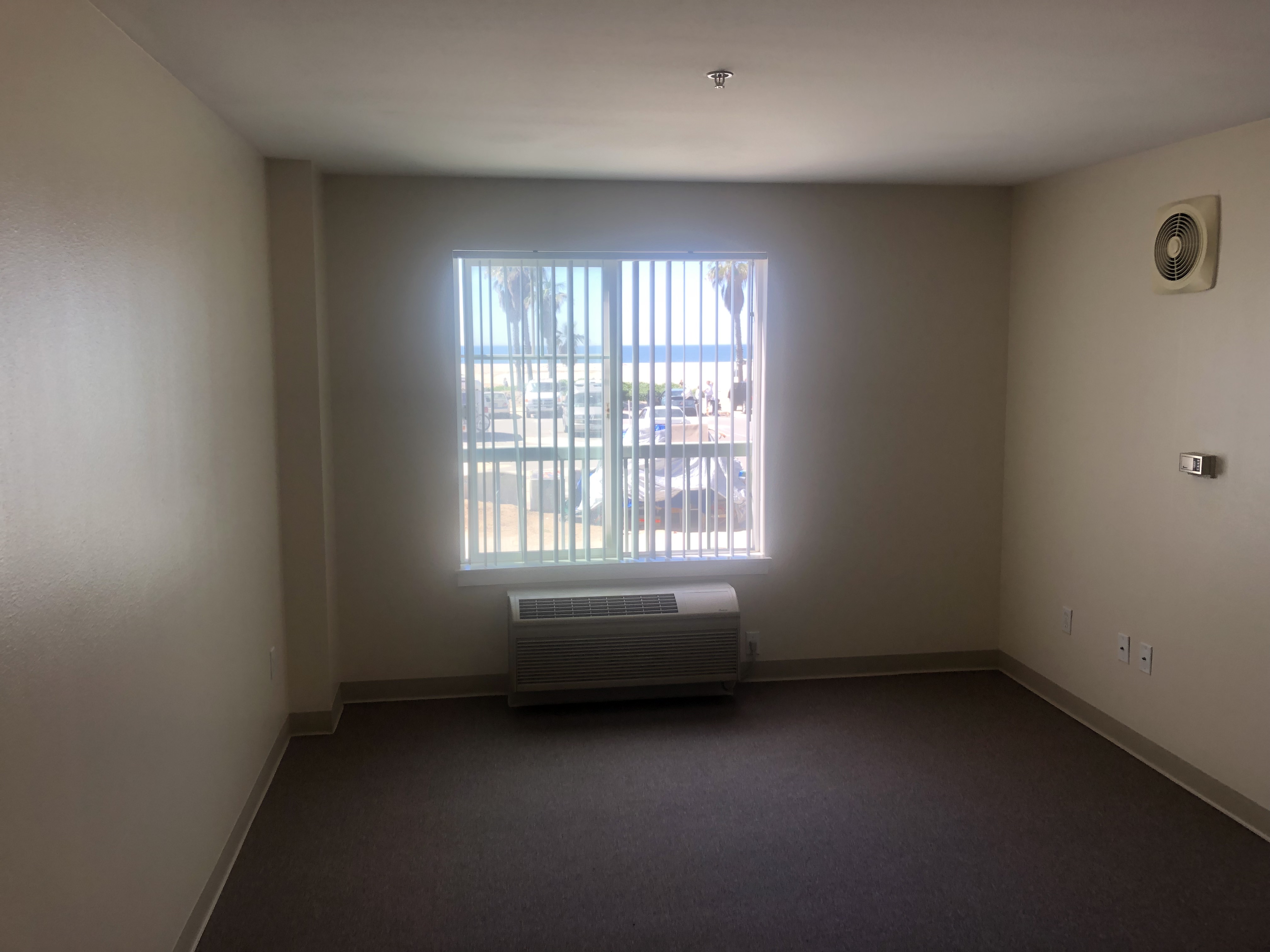 Inside view of an empty bedroom. There is a window with white vertical blinds and an AC below it. On the wall there is a thermometer dial and a spinkler on the ceiling.