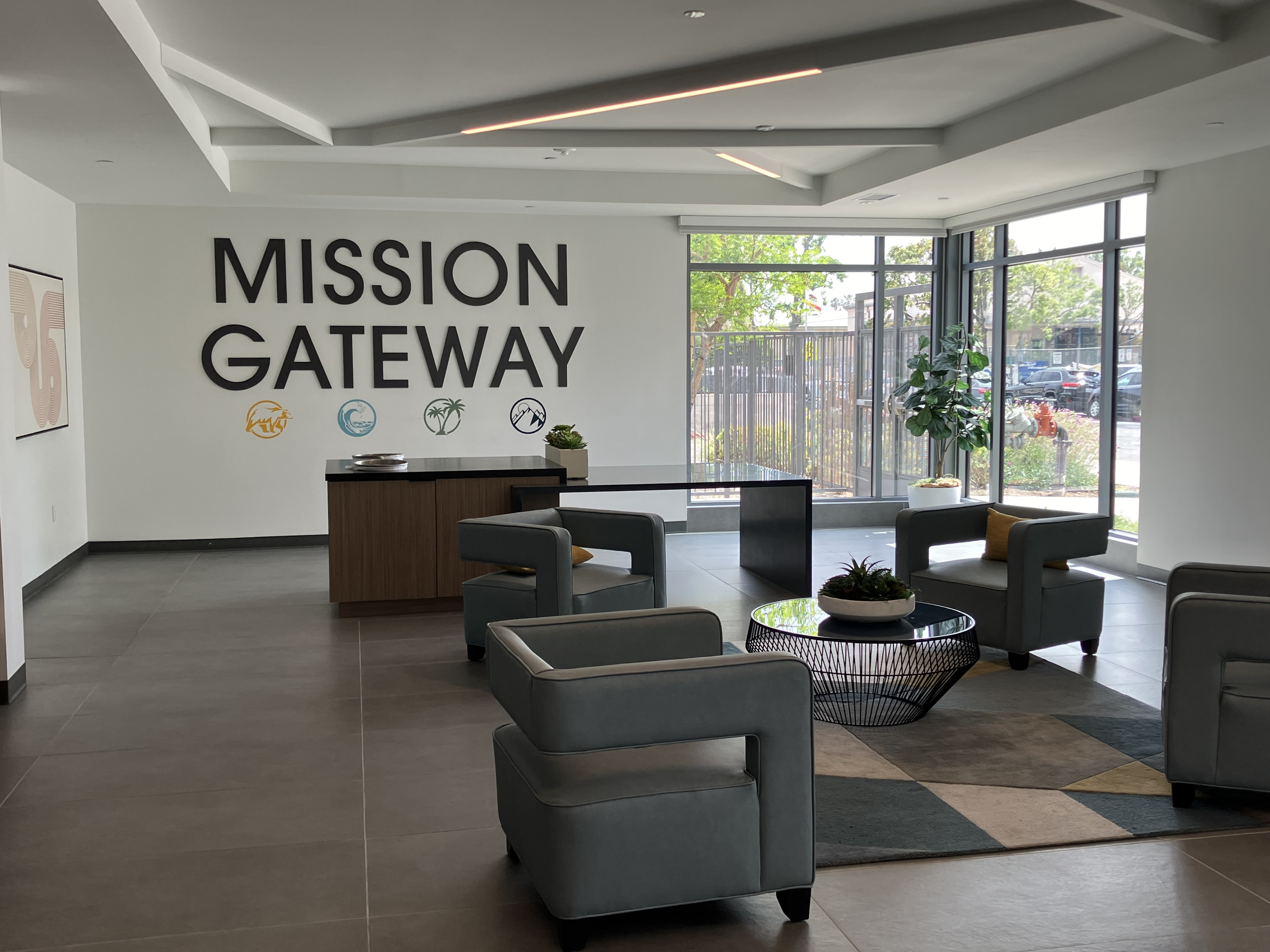 Mission Gateway Lobby Building 1 on Langdon Ave. Entrance to building, there are 4 chairs and a center table, a rug under the center table. There is also an island table and some greenery.