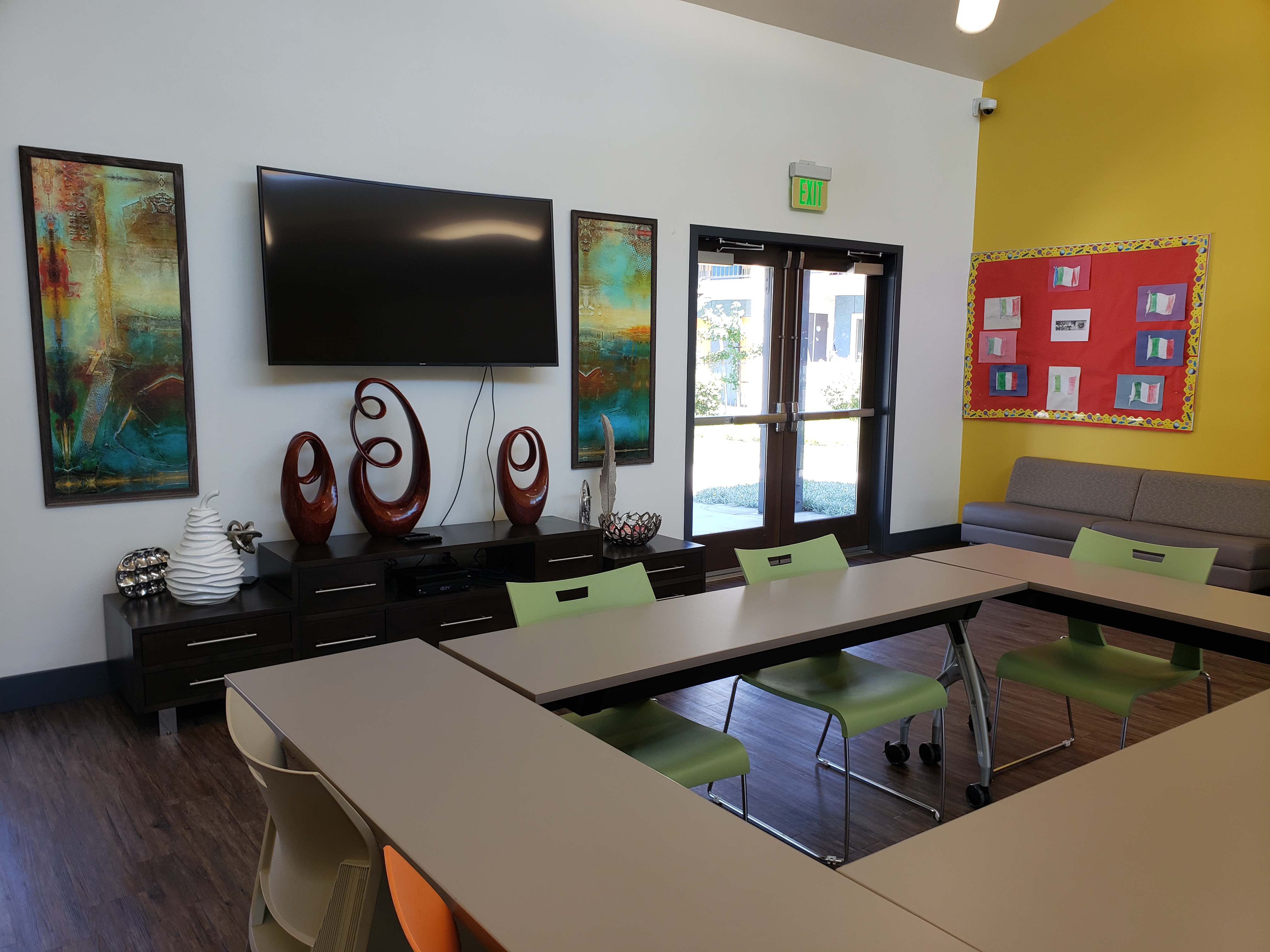 Interior view of a community room with double door ground level entry. Room consists of a small sofa, flat screen tv on the wall, tables and chairs, and modern art decor. Tables are set up to face each other in a rectangular pattern.