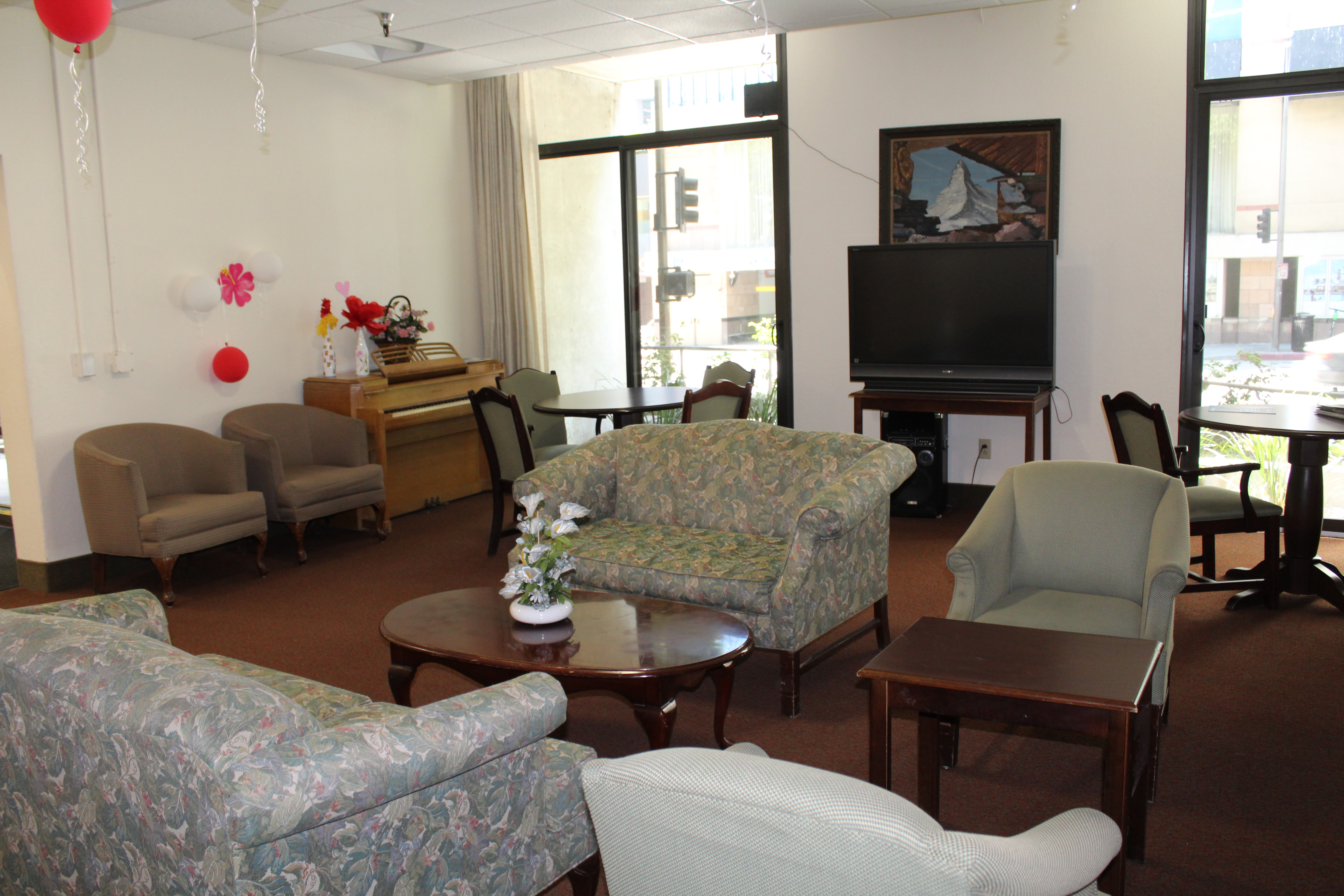 Inside view of a community room. Room contains talll windows with a street view. There is a piano and a tv for entertainment. Furniture consists of sofas, coffee table and two wooden tables with matching chairs.