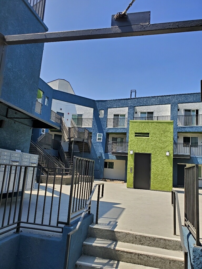 Picture of an entrance way toward a white and blue three story building. There is a small stairway with handle bars on the side leading up to the platform. There is a green colored elavator, multiple staircases, and mailboxes.