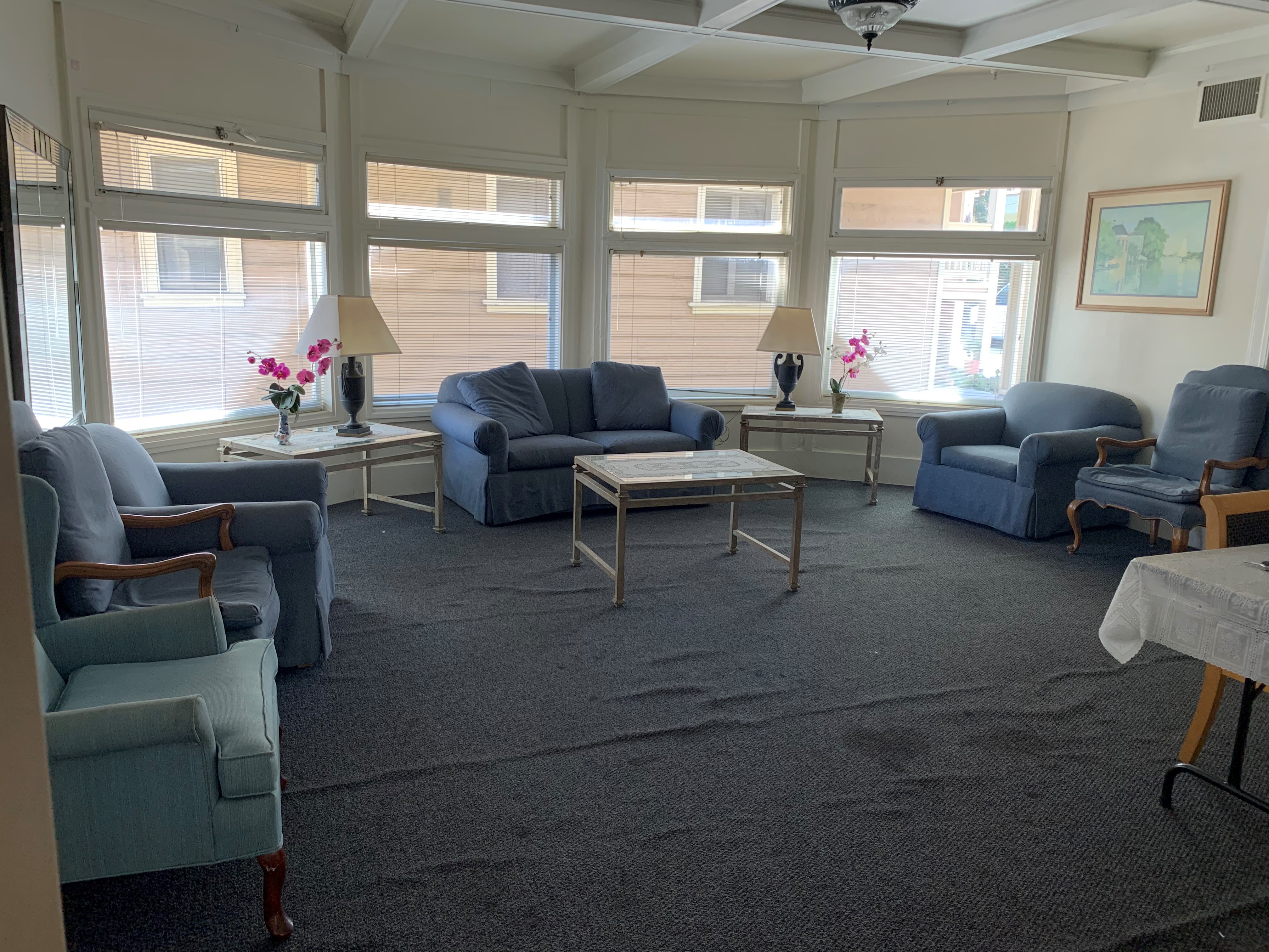 Lounging area with with multiple sofa chairs, a coffee table and side tables with lamps and plants. Floor is carpeted. There are four windows with blinds.