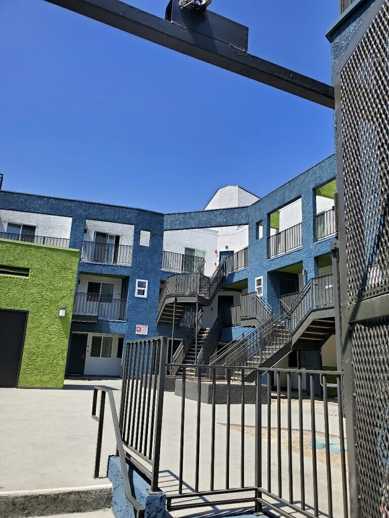 Front angle view of green elevator. Behind it is a three story white and blue building with a flight of stairs.