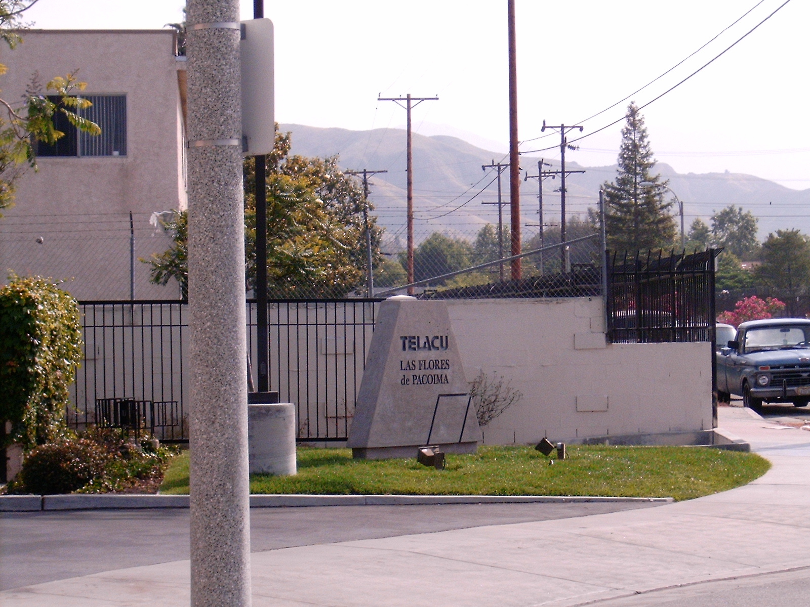 Corner view of the building, concrete standing stone with the building's name "Telacu Las Flores de Pacoima", old blue parked truck, grass and plants in front of the gated building.
