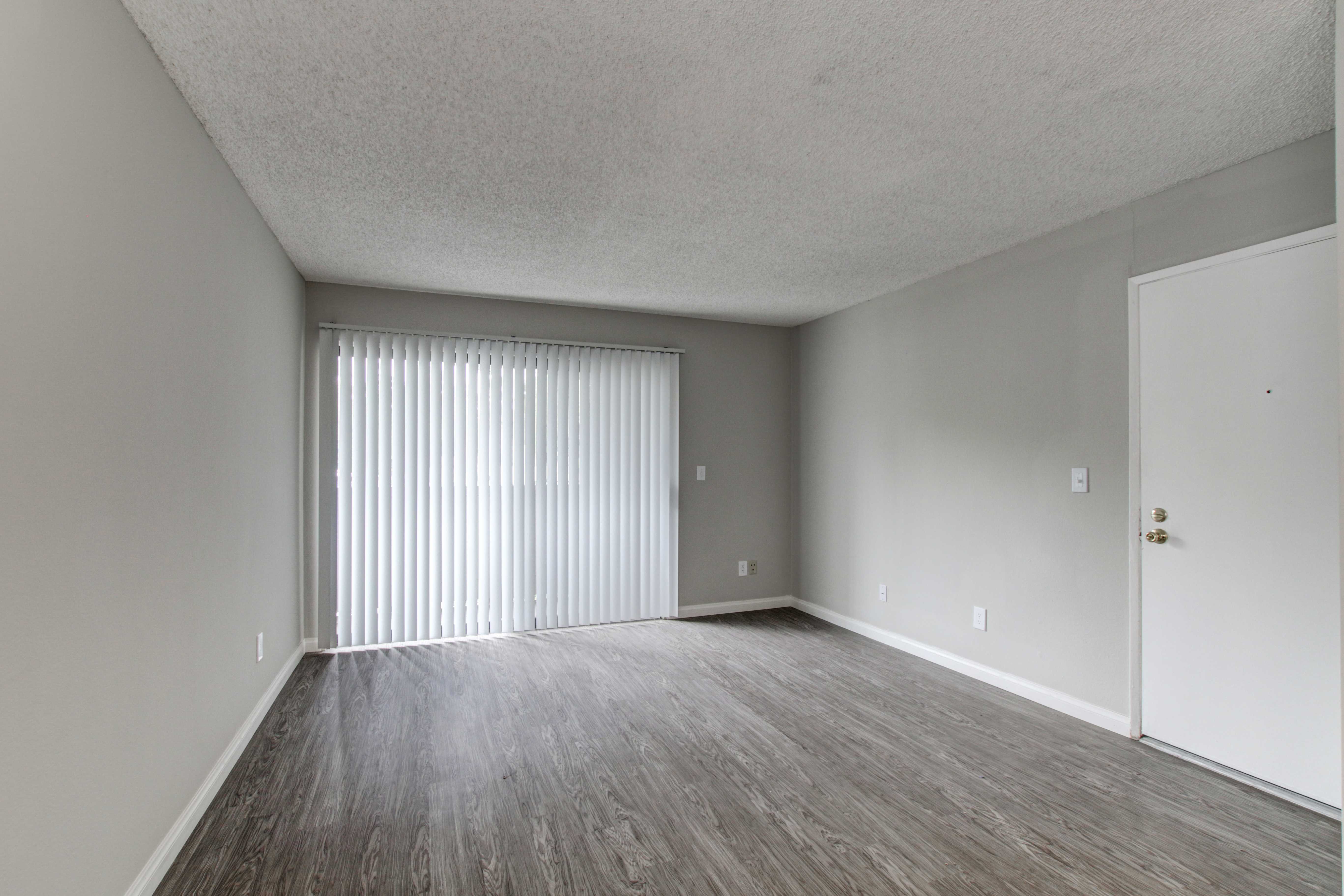Inside space of a unit. Visible is a large window with vertical blinds, door, multiple outlets, and wooden floors.