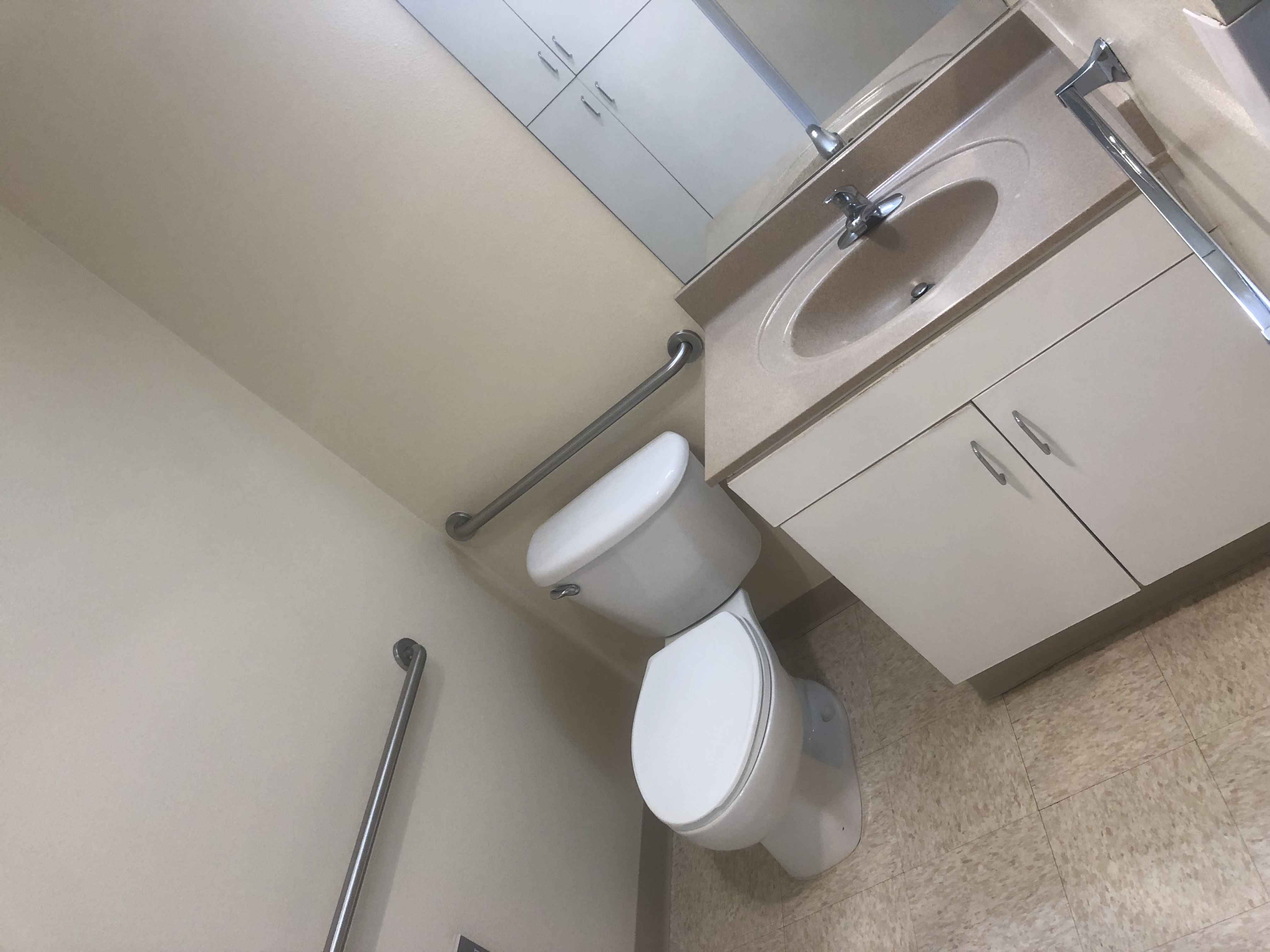 Inside view of a restroom that consists of toilet, sink, mirror and handle bars. There are cabinets under the sink and across the room.