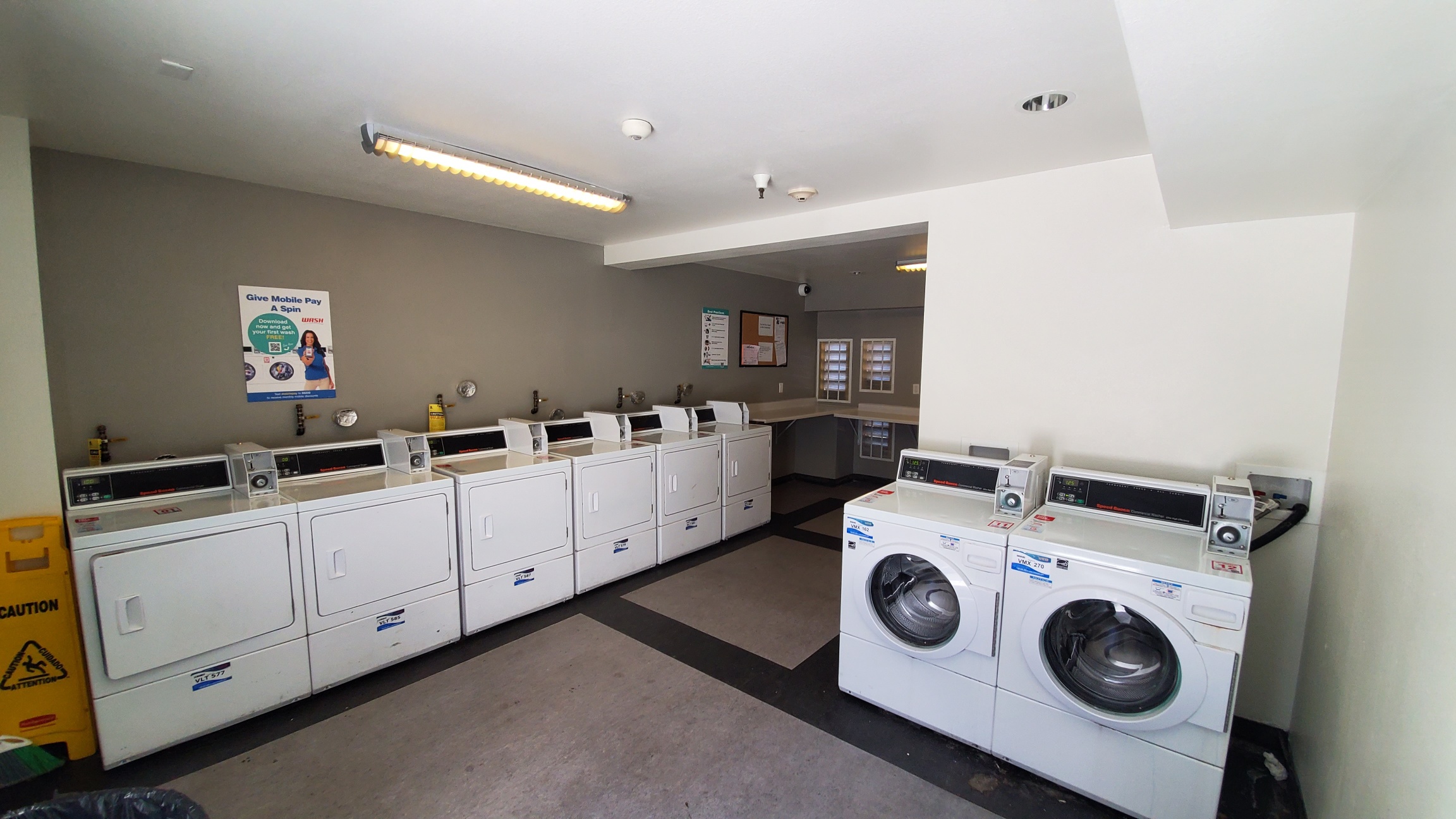 Different angle of laundry room with multiple washer and dryer machines. Room has a shelf for folding that is L shapes along the walls. There is a security camera on an upper corner of the room, and posted signs across the walls.