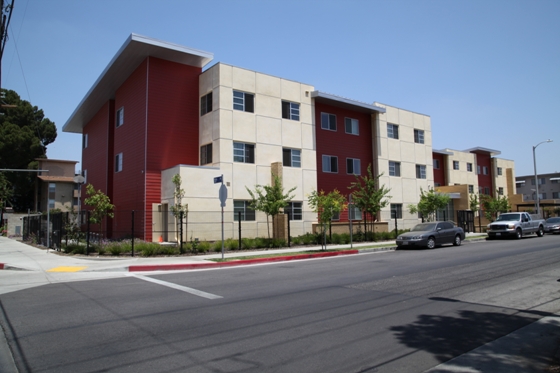 Image of white and red 3 floor building, with green plants and small trees planted along building, and street parking in front of the building