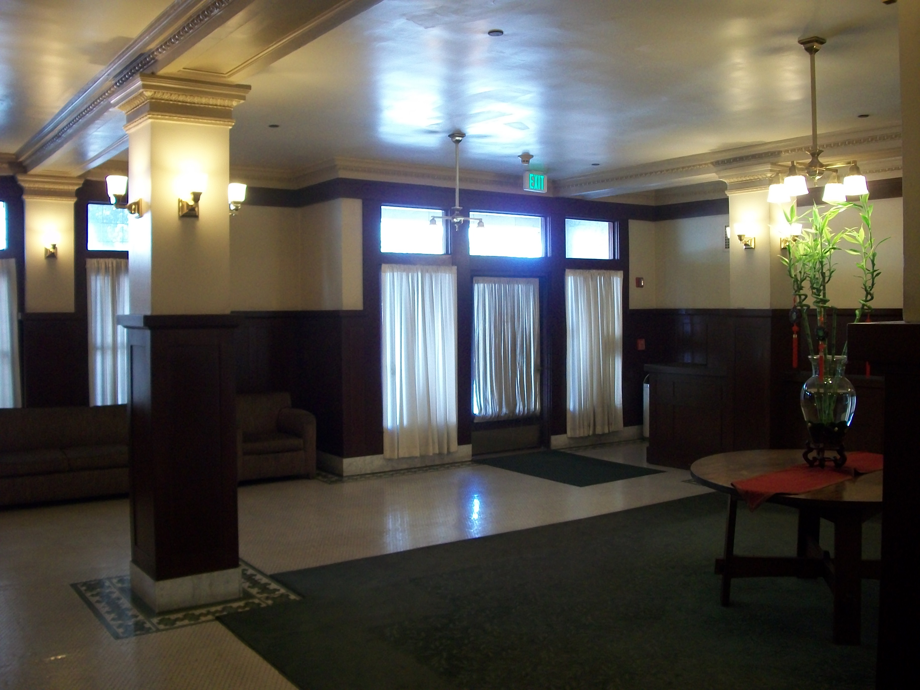 Interior view of the Lobby area of the Young Apartments. Glass door and windows coverd by a thin whitecurtain. White and brown pillar with a carpet in the middle with a table in center