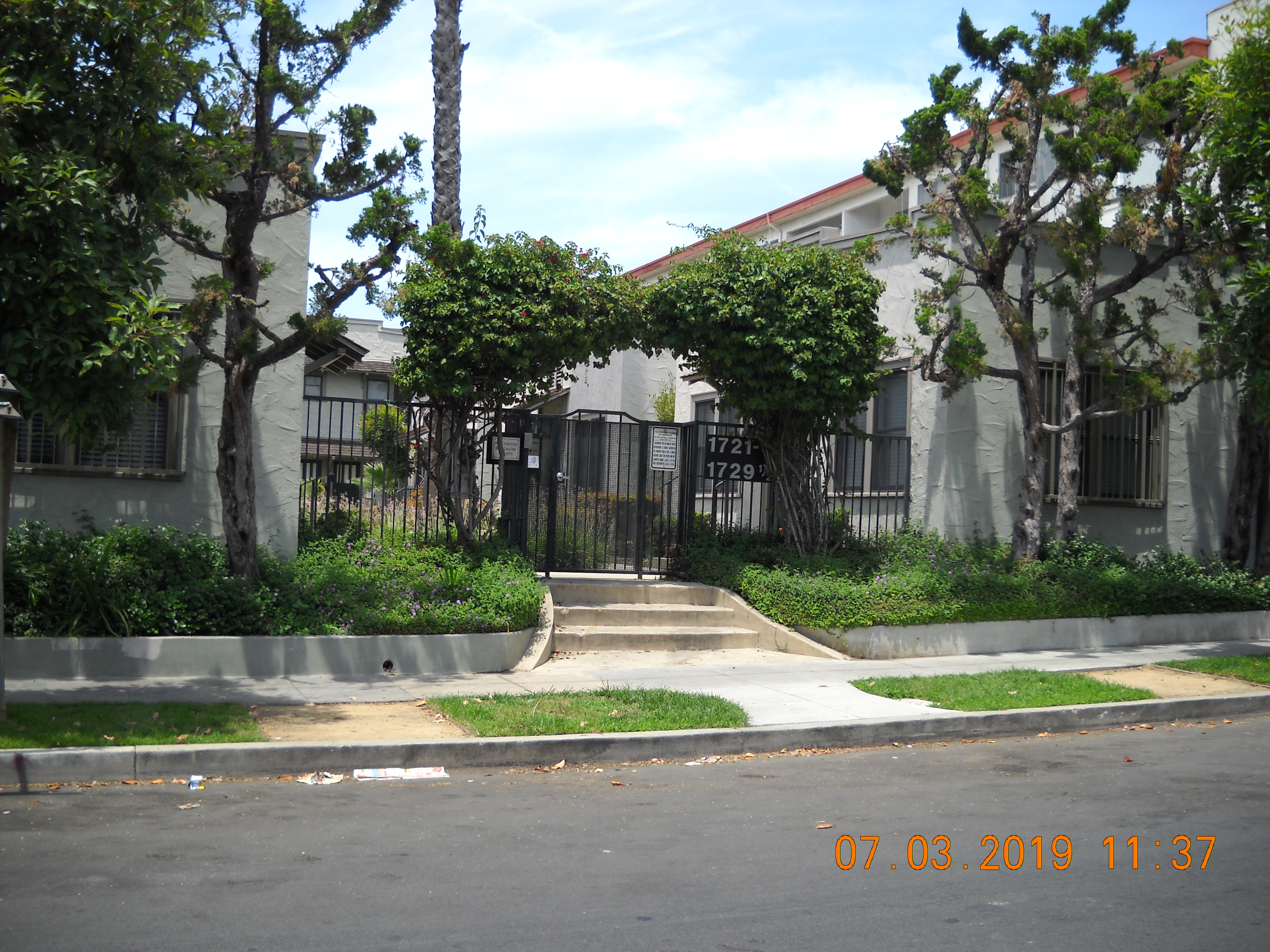 Front view of a two story gray building complex. Entry is gated, has a keypad, and three steps to enter. Units wrap around a center that contains plants, trees and bushes. There are trees and bushes in front of the building as well.