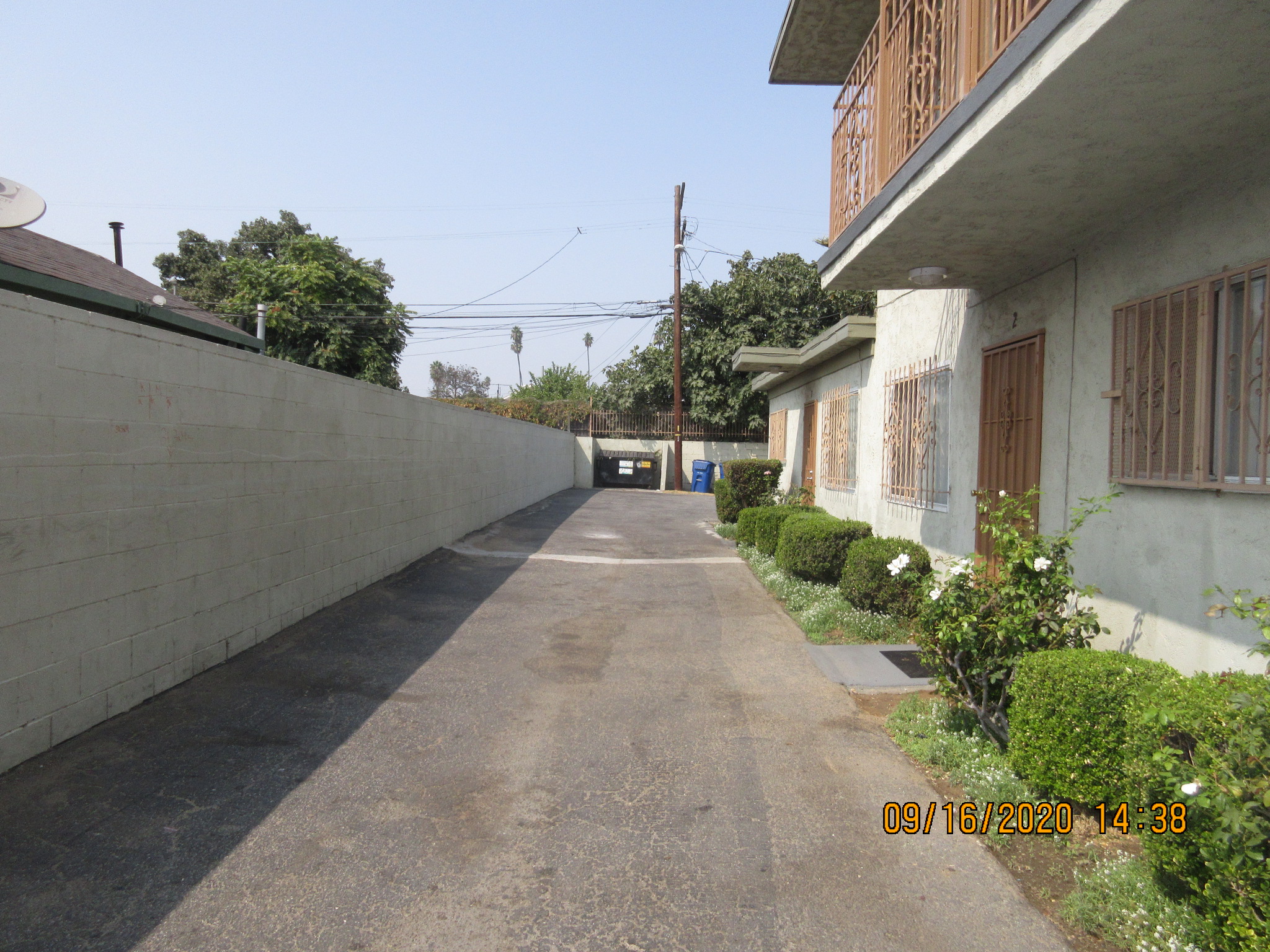 Windward A&B exterior view. Long paved driveway in front of building with trash bins at the end. bars on the window