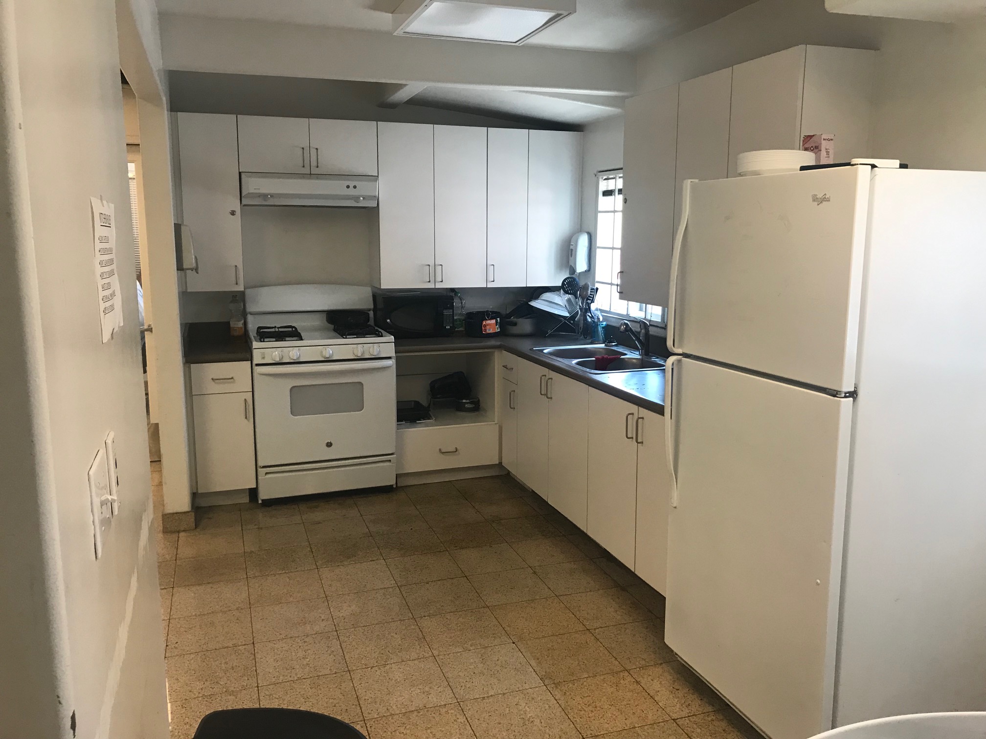 Front side view of a kitchen, white pantry, large cabinets, white stove with oven and under cabinet range hood, black microwave, pots, dishes, gallon of water, coffee maker, soap dispenser, sink, white window, white refrigerator, ceiling lights, and tiled floor.