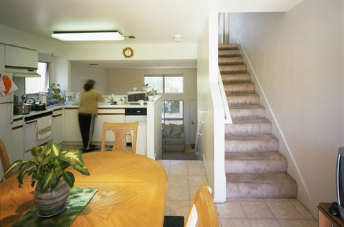 Interior view of a multi-level unit at La Brea/Franklin Apartments. A women at the kitchen sink with a table and chairs behind her with stairs leading up to the side of her.