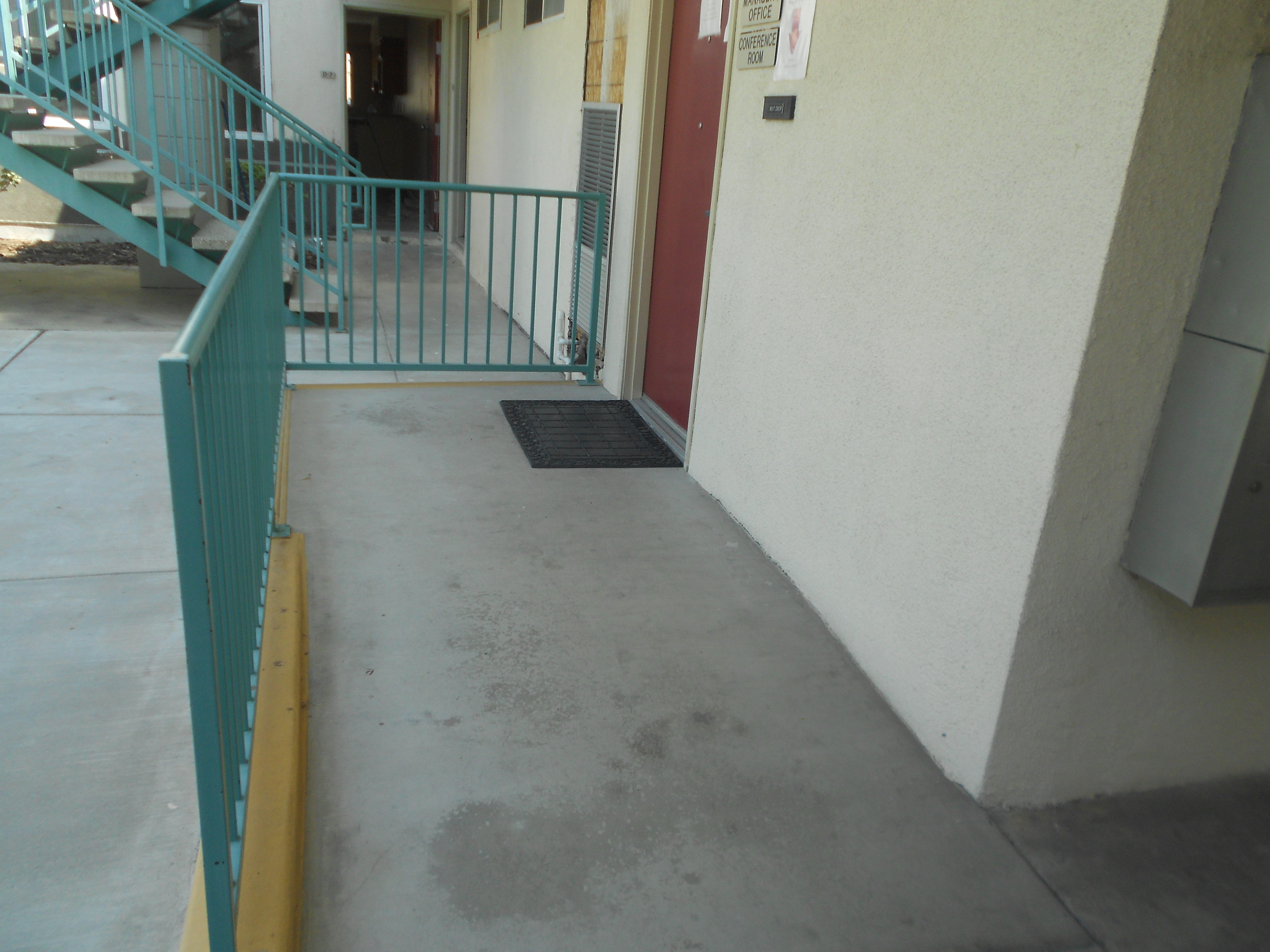 View of a ramp that leads up to conference room with a maroon door. Ramp has a turquoise railing on the side. There is a staircase with handlebars that leads upward in the back.