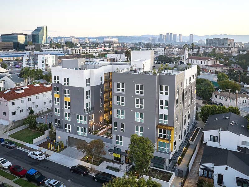 A modern mid-rise apartment building surrounded by residential homes, with a downtown LA skyline visible in the background.