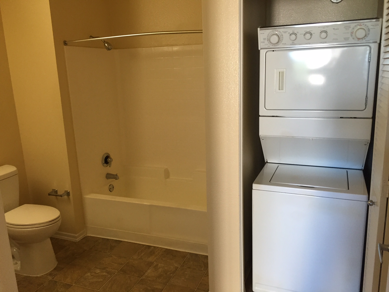 Front view of a bathroom, white toilet and white tub, shower rod without a curtain, empty toilet paper holder, a compact white two in one washer and dryer machine on the right.