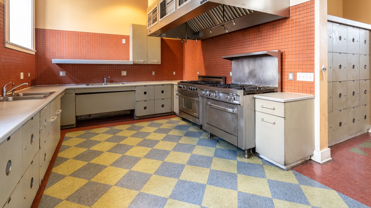 Commercial type kitchen, two stainless steel stoves, stainless steel range hood, white and beige pantry, two sinks, blue and yellow diamond tile floors.