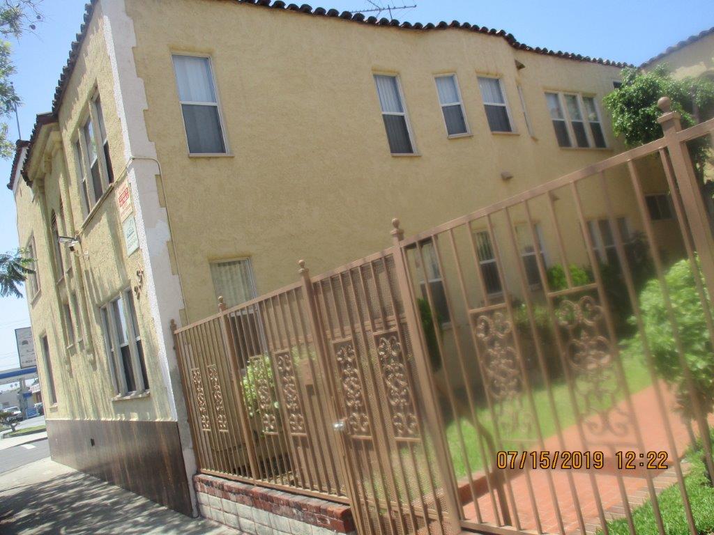 Corner side view of a yellow two story building, locked gate entrance, brick entrance, multiple trees and bushes on each side of the entrance behind the gate, cameras and no public and management company signs.
