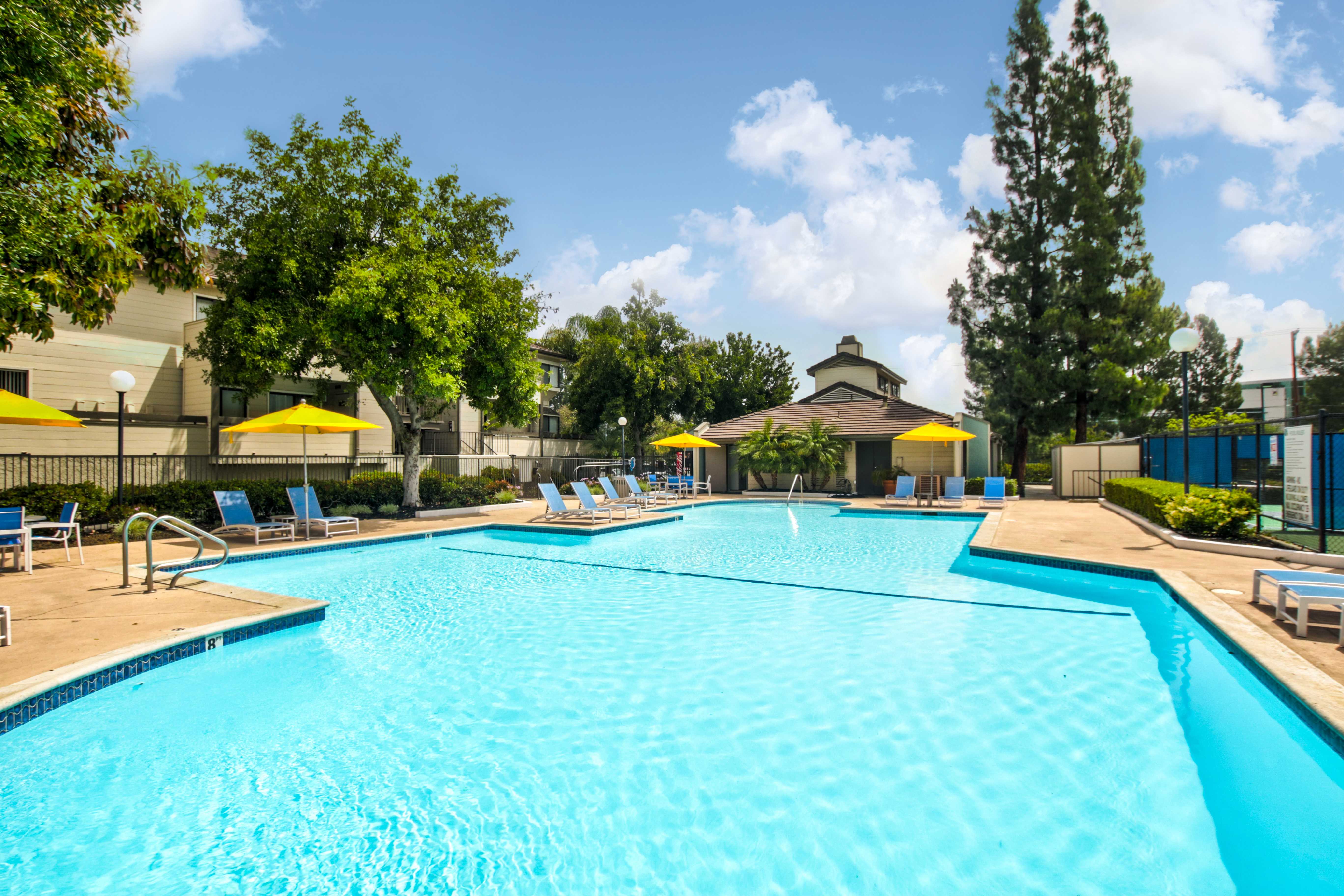 Different angle of pool that is surrounded by multiple lounging chairs, small circular tables, and yellow umbrellas. Across from it is a small hut. Plants and trees surround the perimeter of the area.