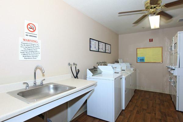 Side view of a laundry room, a sink and four white washer on the left side, beige walls and white ceiling, ceiling fan, four white dryers on the right side, a board with flyers, no smoking and warnings signs on the left side wall.