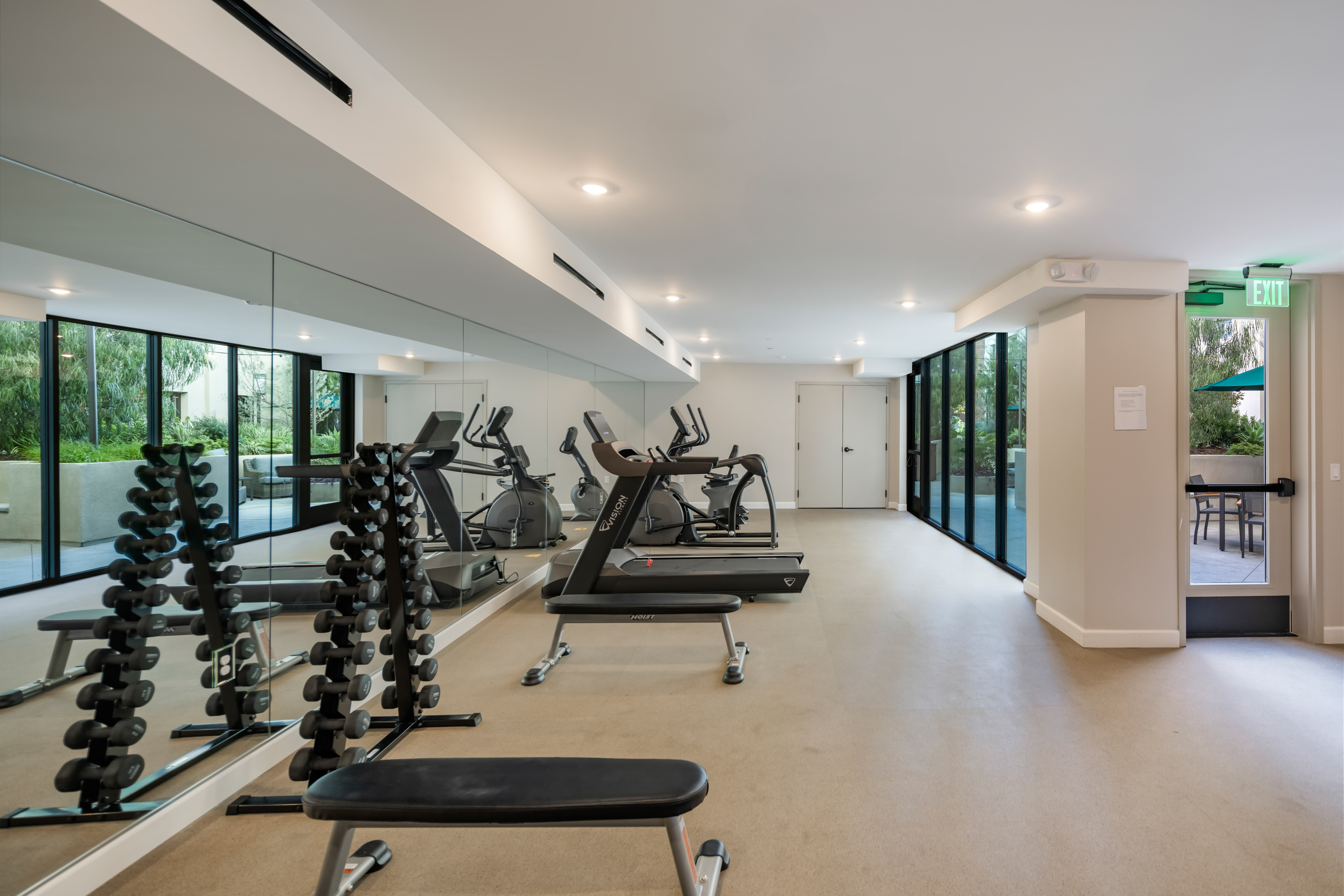 Modern gym with mirrored wall, treadmills, stationary bikes, and dumbbells. Bright, clean space with large windows and a tranquil outdoor view.