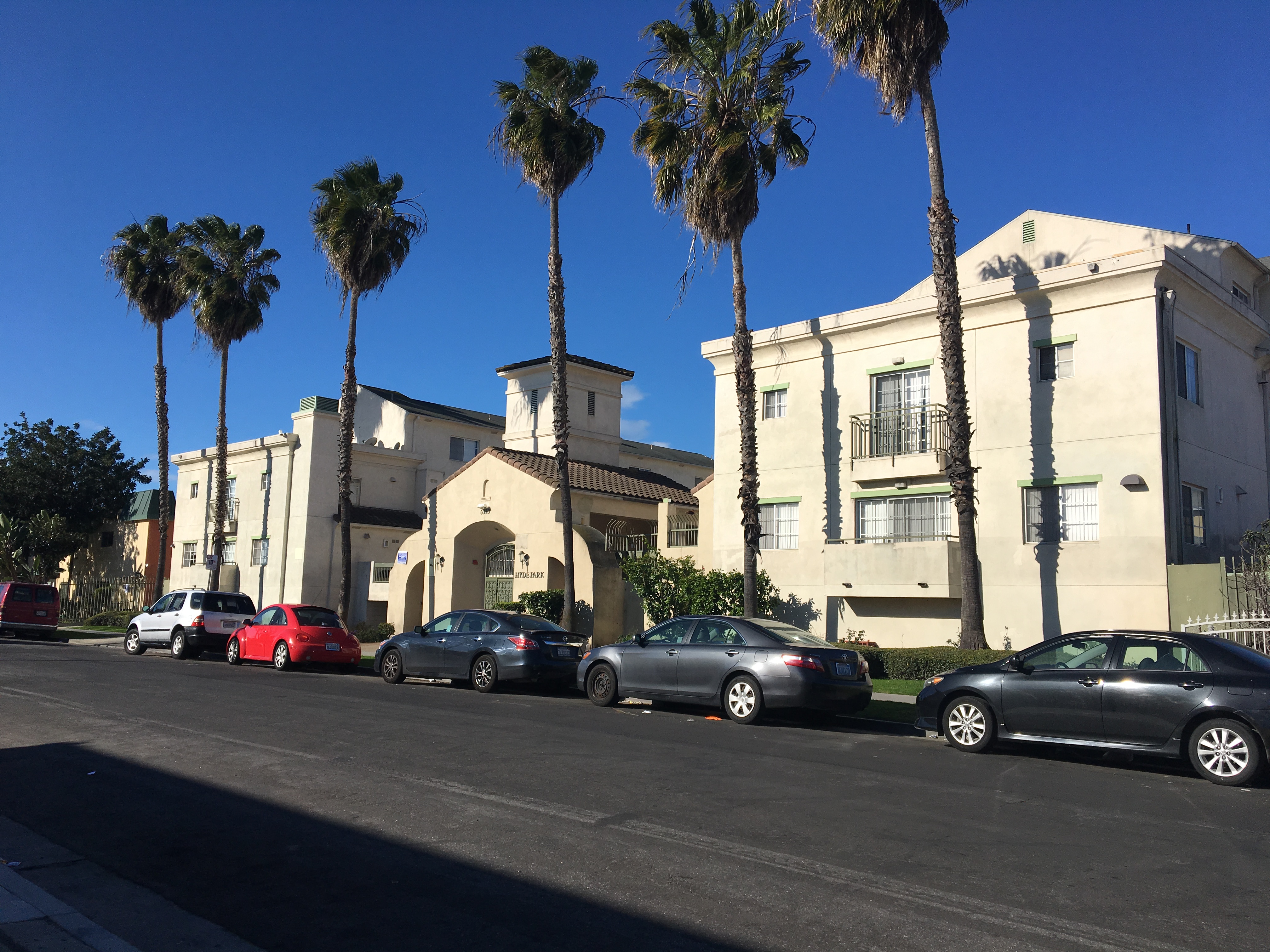 Street view of Hyde Park Place Apartments. Palm trees line the sidwalk along the building with cars parked on the street in front of the building