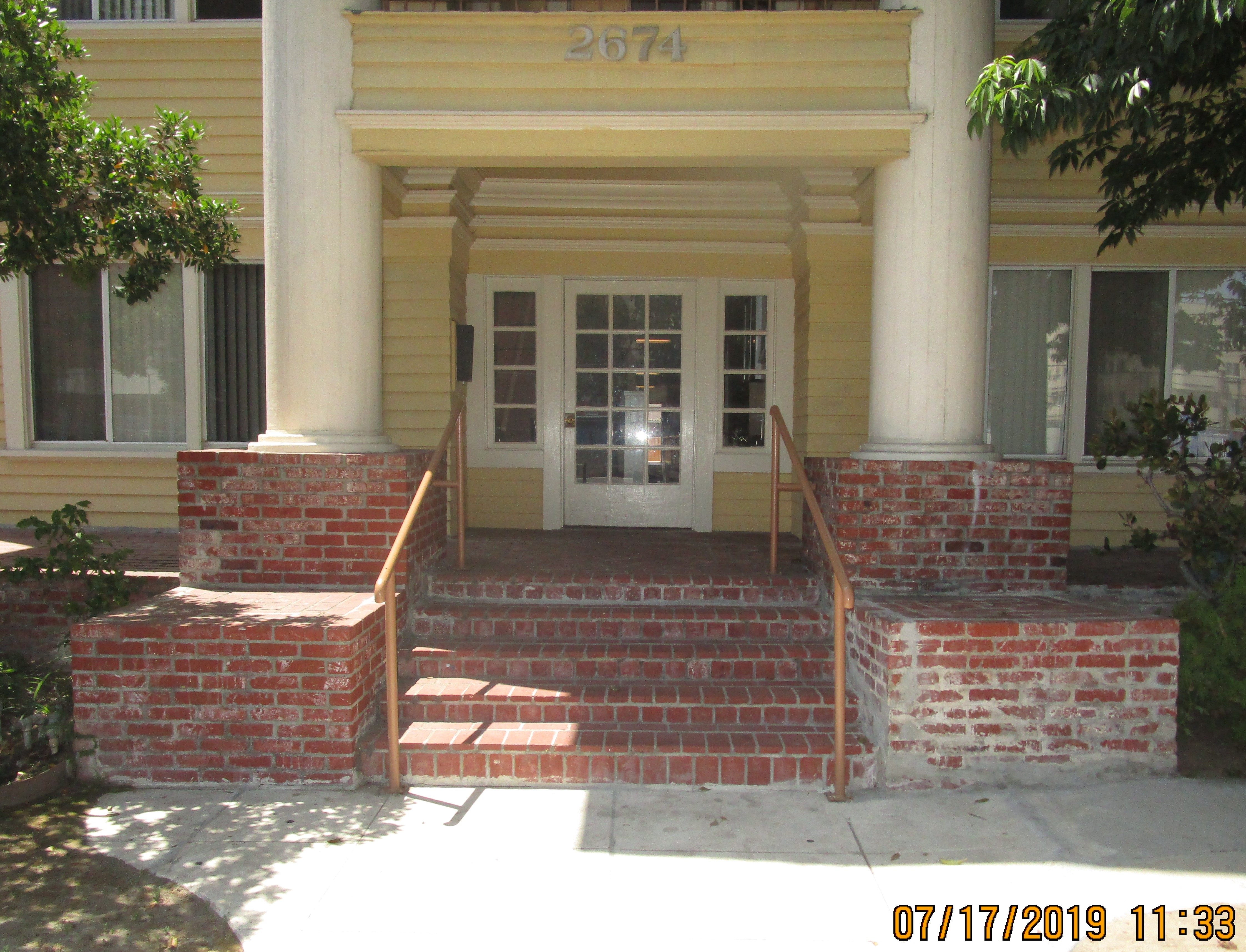 Brick paved front entrance and porch of yellow building with hand rails and two white columns on each side of the steps.