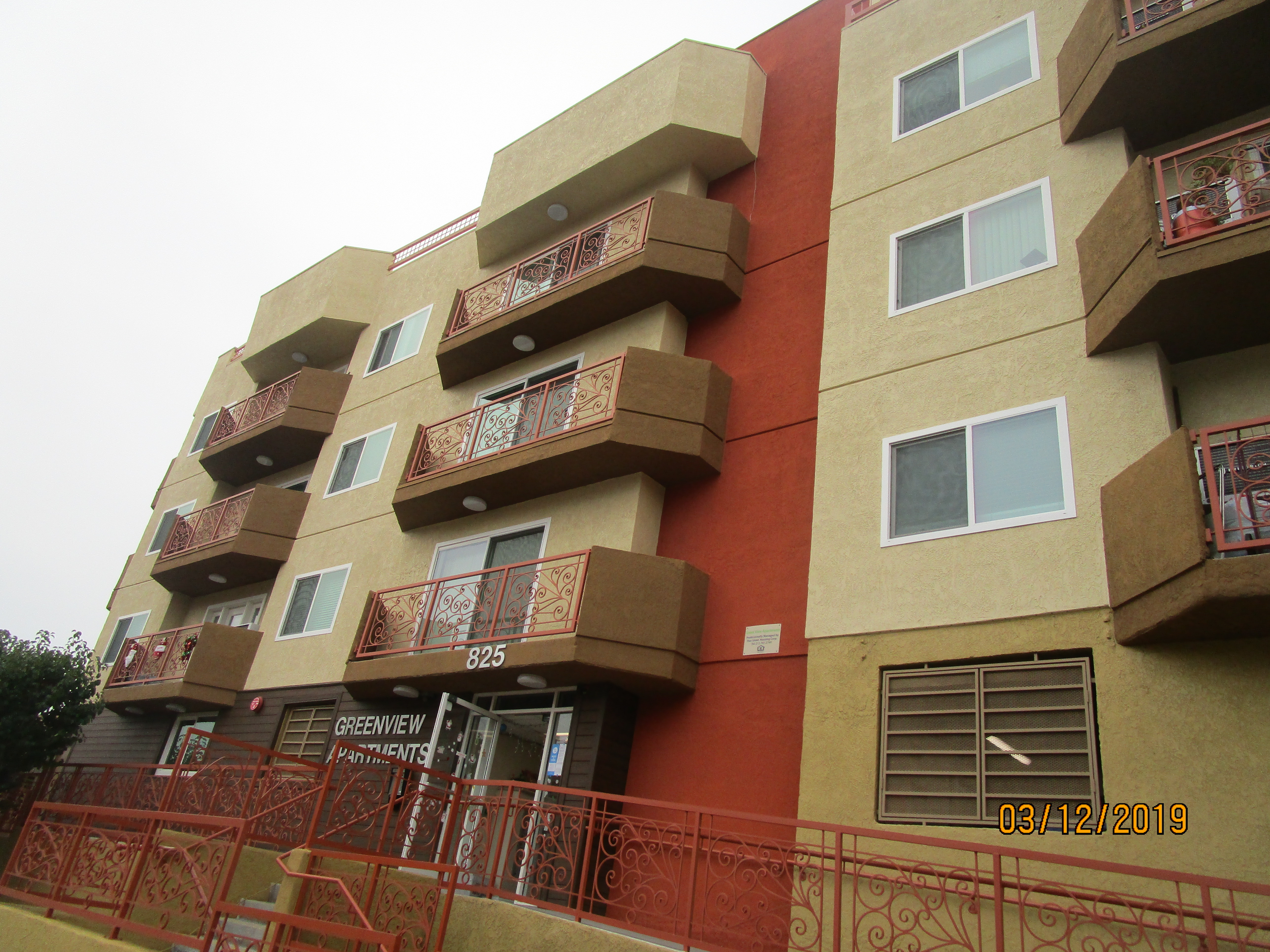 Right side view of a colorful four story building, multiple windows, some windows with balconies, orange metal hald way fence with handrails, steps and a ramp.