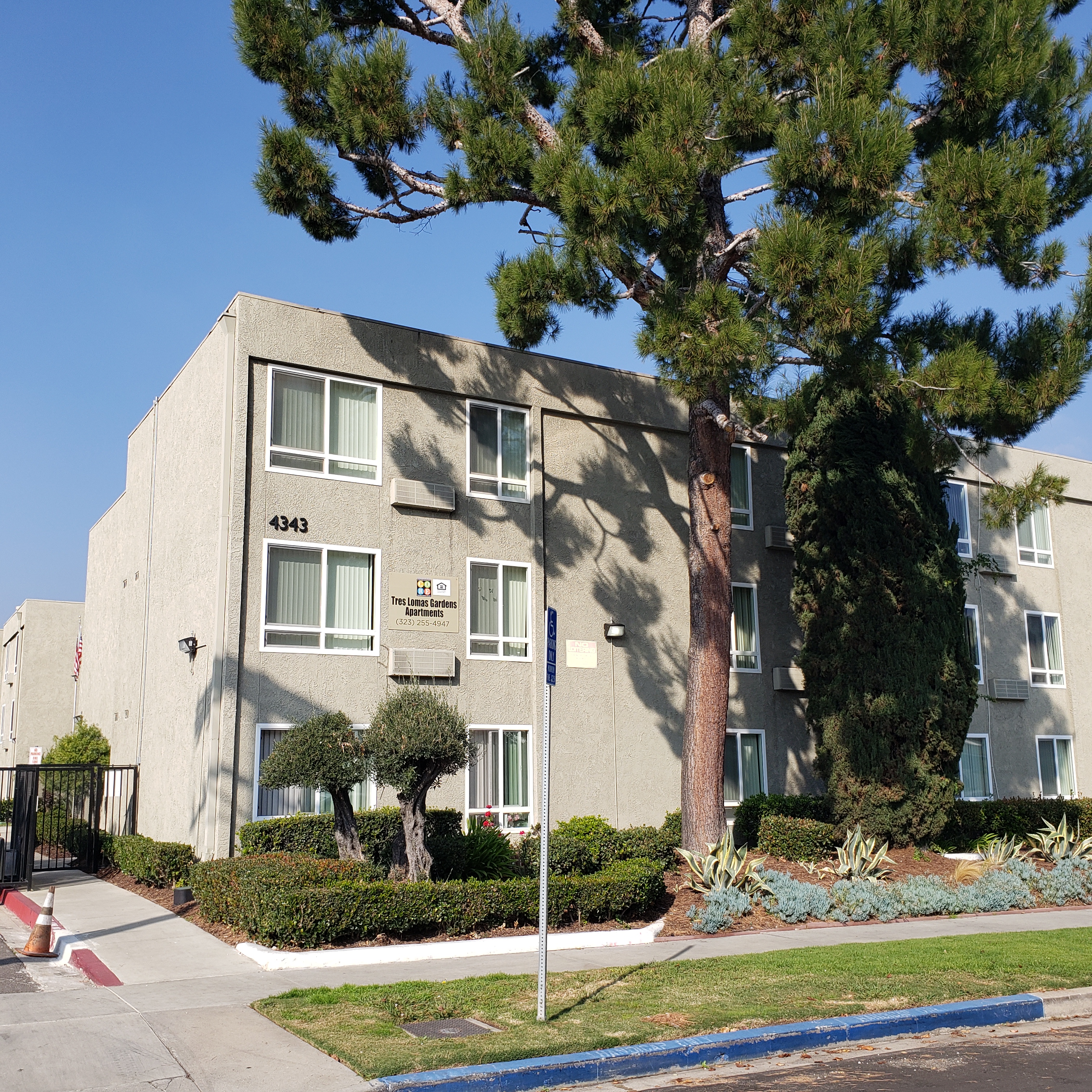 Front view of tres lomas garden apartments. multi-story building with large trees, bushes and grass in the front. Section of curb on street is painted blue with parking accessibility sign.