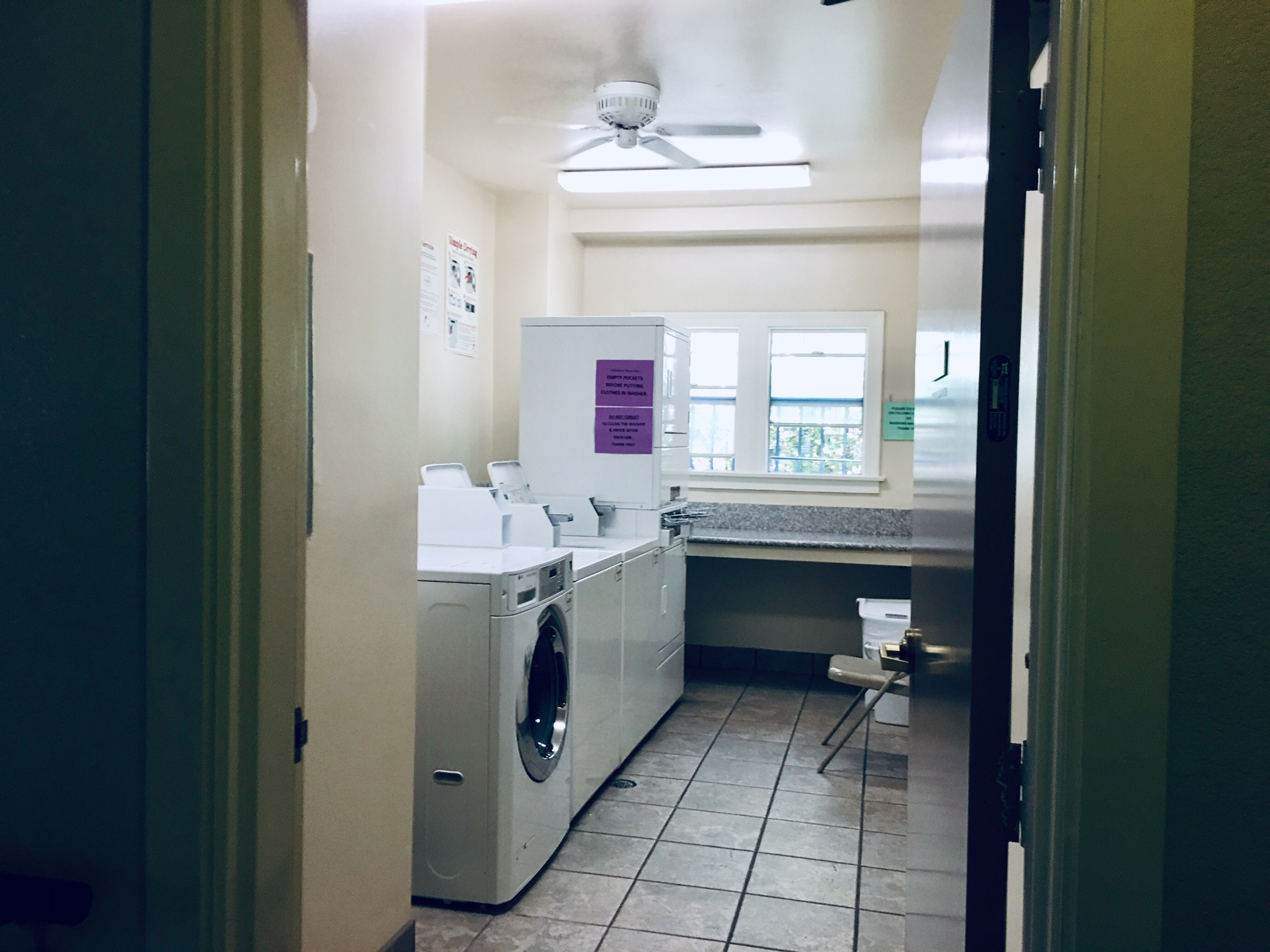 Laundry room with multiple washers and dryers. There is a folding shelf under the windows, and posted signs across the room. There is also a folding chair and a trash bin inside. Room also has a ceiling fan.