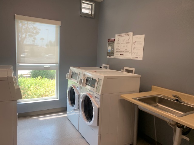 View of a laundry room, white washers and dryers, signs posted on the wall, white window with white shade, a sink.