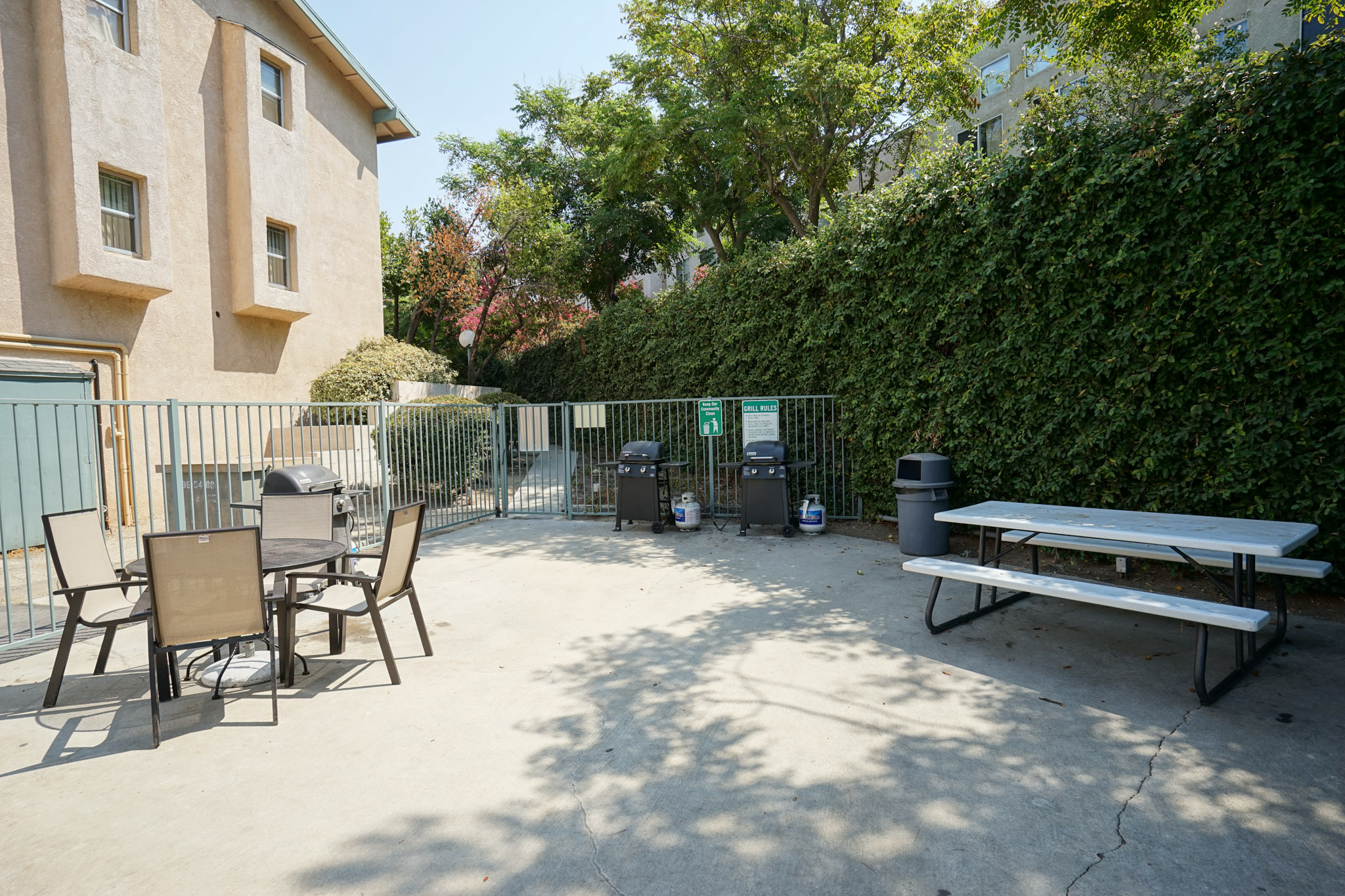 View of a fenced patio area with a grills and two table sets. On one side of the area there is a hedge, and the other side shows a three story building.