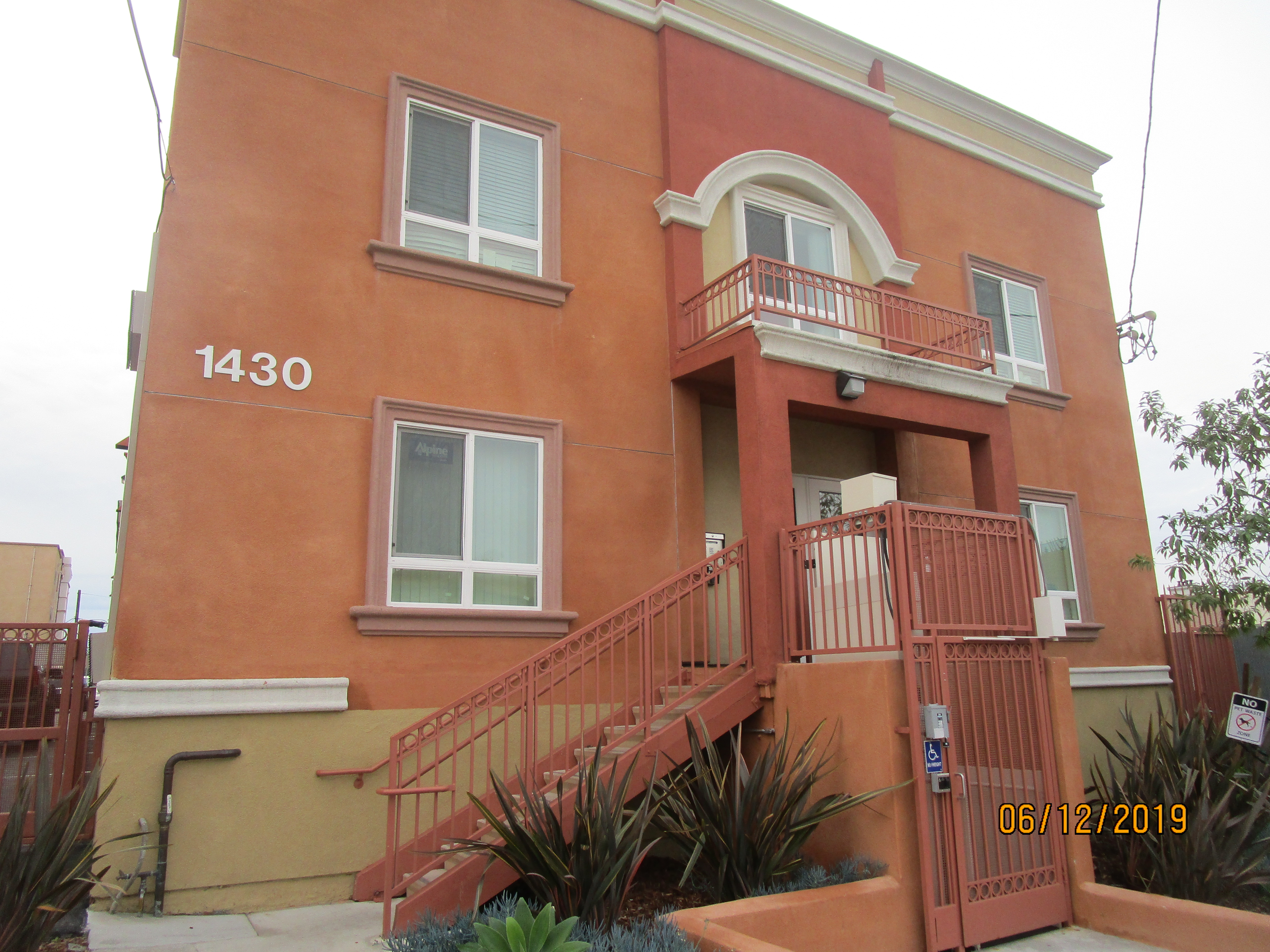 Side view of a colorful two story building entrance, steps with a handrail on one side, a lock door entrance to a ramp, five windows, a middle window with a balcony, bushes and plants.