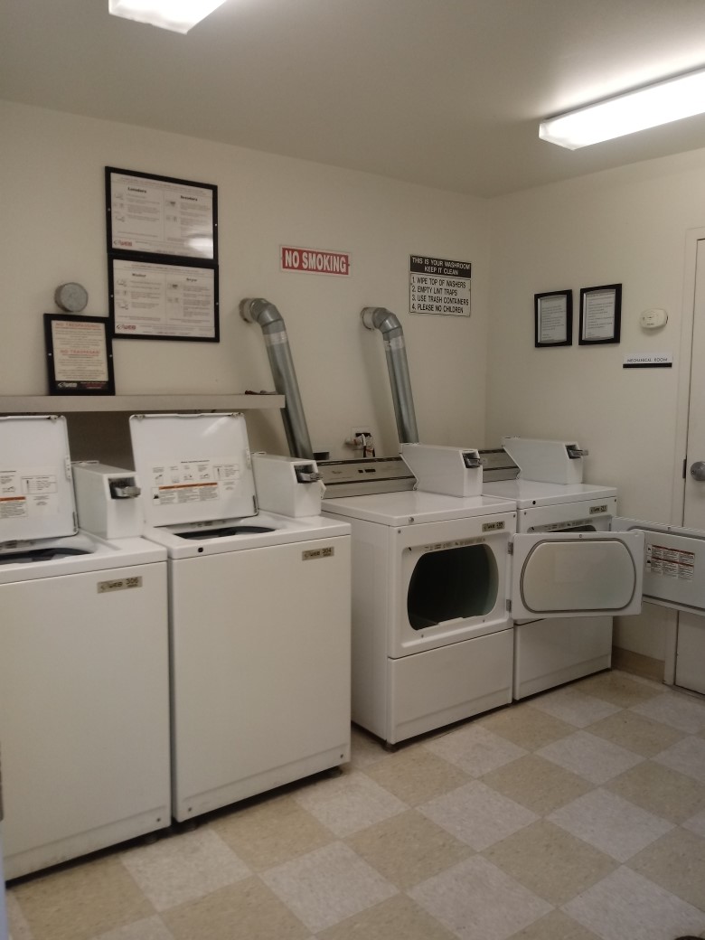 Alexandria House laundry room. Image shows two top load washers and two front load dryes next to each other along wall. Notices and signs hung on wall over washer and dryers.