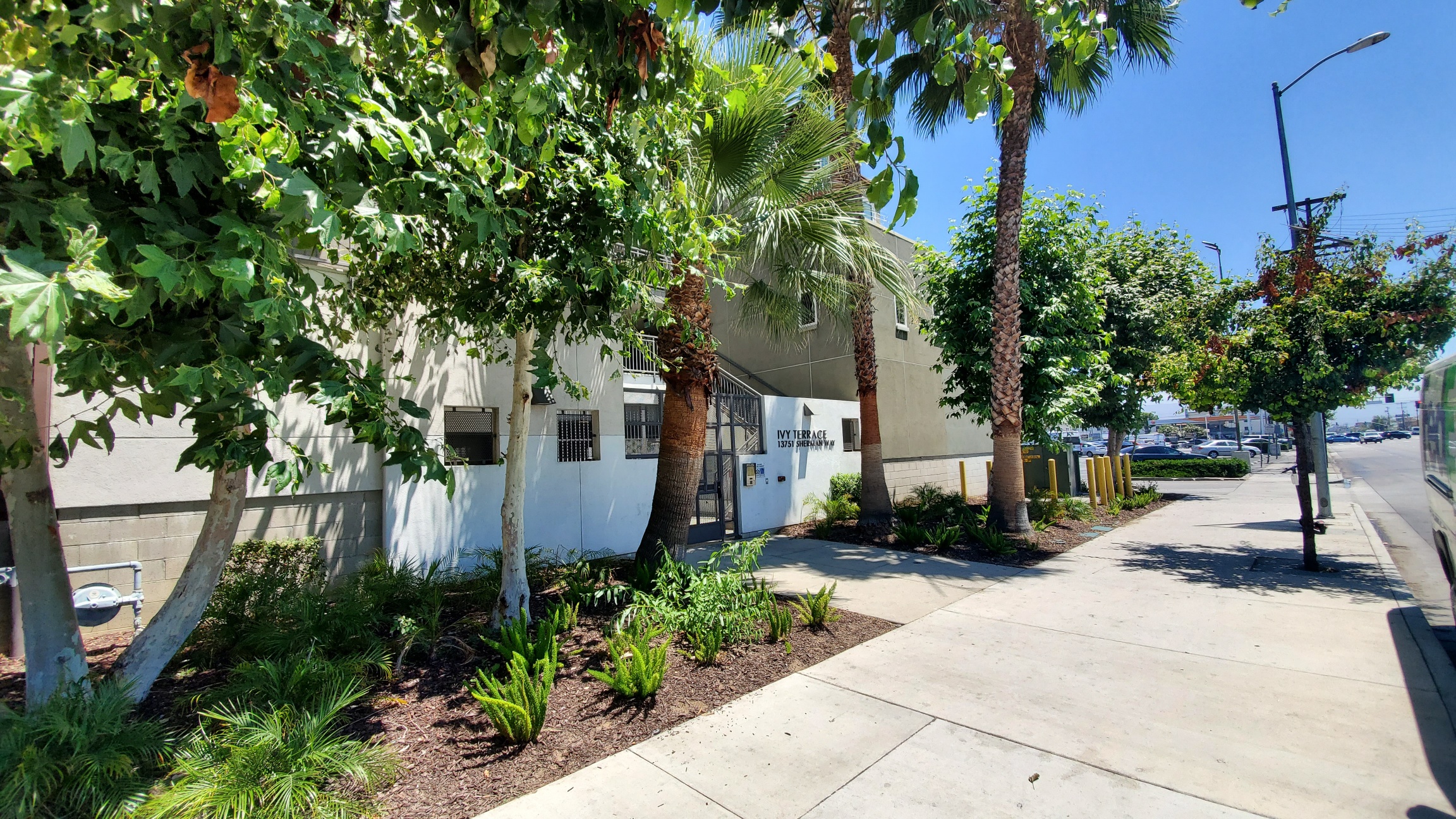 Front side view of a building complex. There is a small front lawn in front of the building that has a variety of plants and trees. Front of the building is gated and has a keypad for access. There is a upward staircase viewable with handle bars on both s