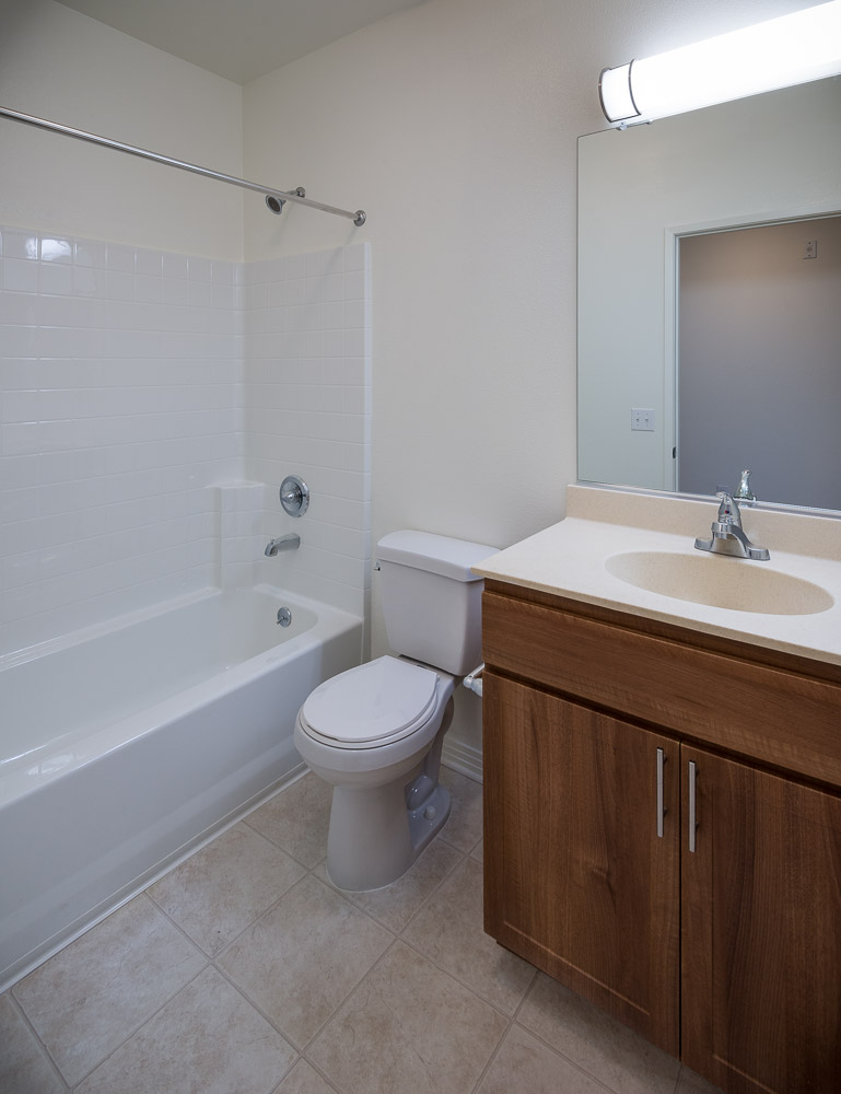 Inside vew of a unit restroom in Rio Vista Apartments. Tile floor with white bathtub, toilet and brown cabinets with a large mirror above.