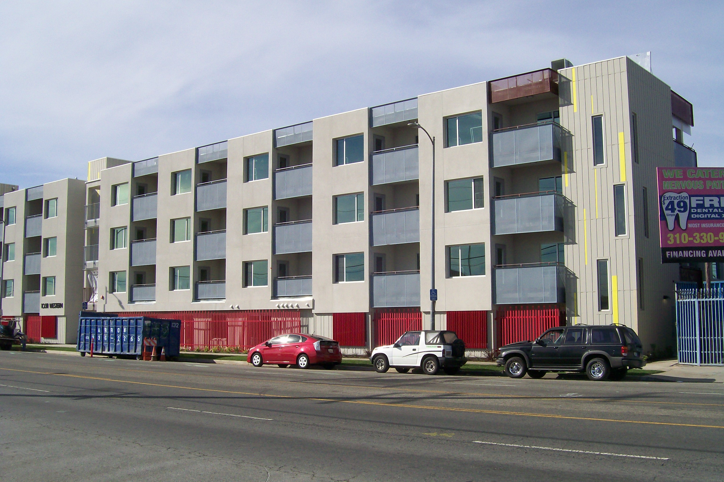 Side view of a modern colorful three story building,red gate, multiple windows, parked cars, big blue portable storage container with orange traffic cones, a handicap parking sign, light pole.